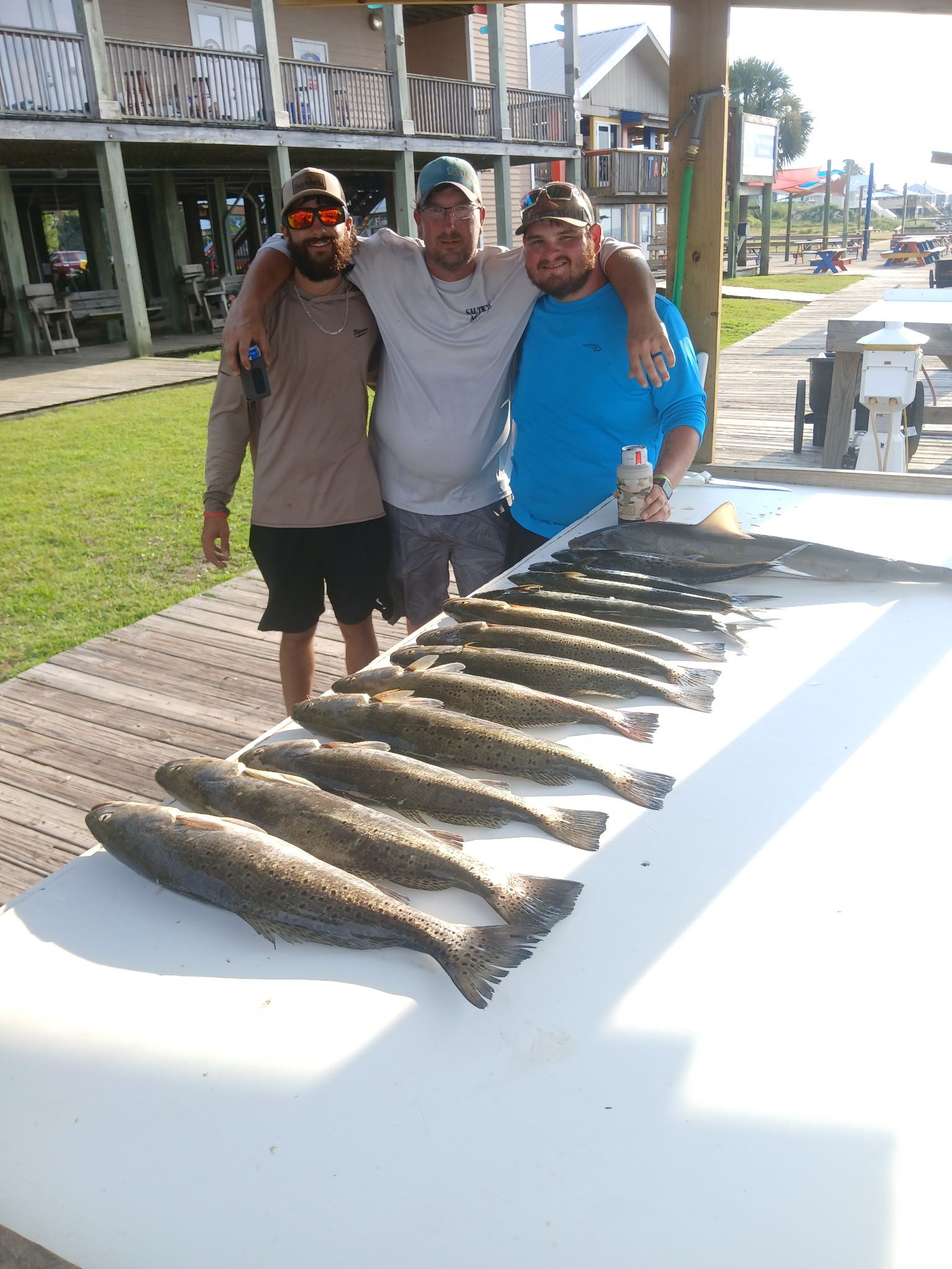 Three people stand behind a table lined with fish outdoors on a sunny dock.