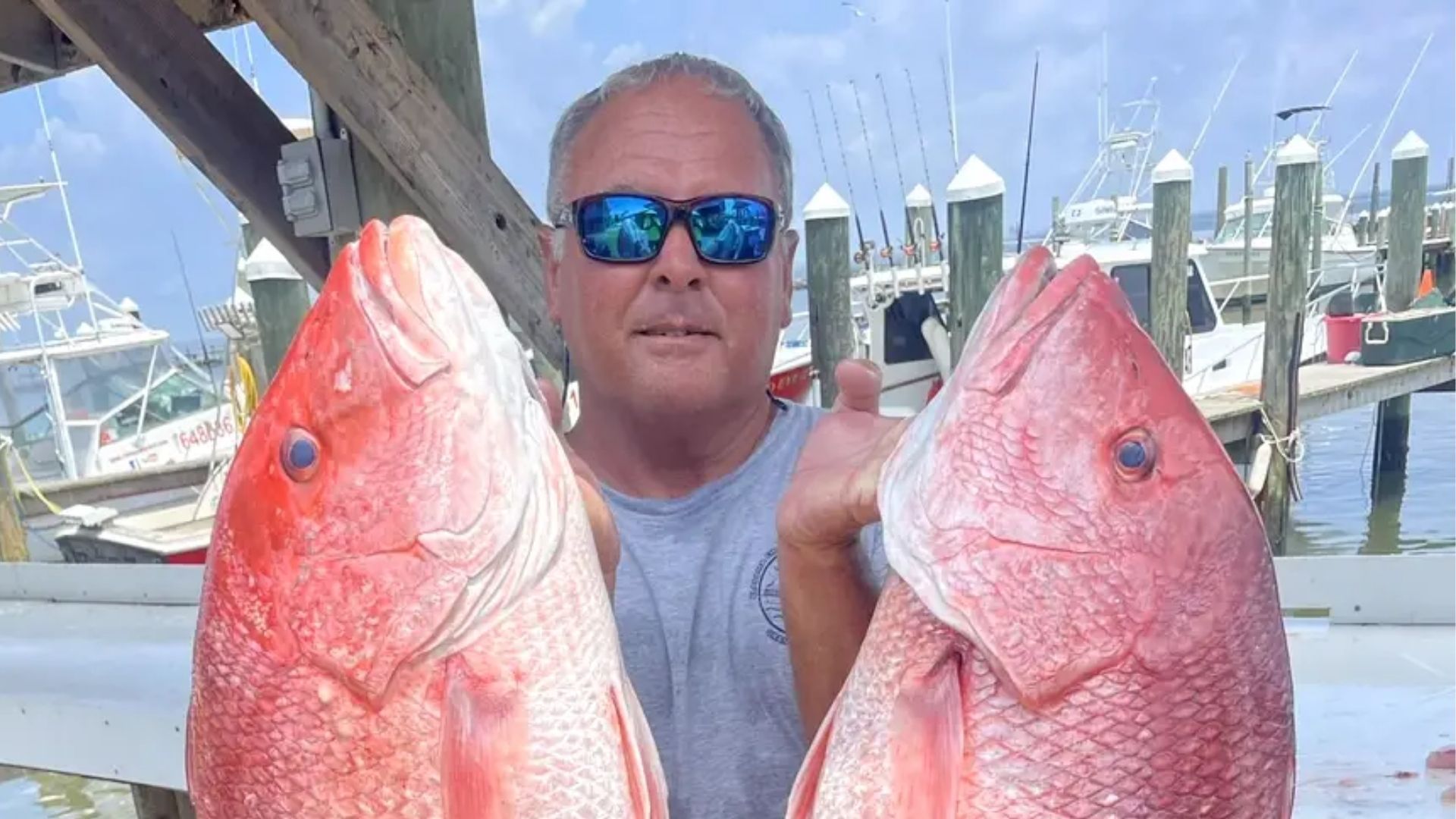 A person holding two large red snapper fish at a marina with boats in the background.