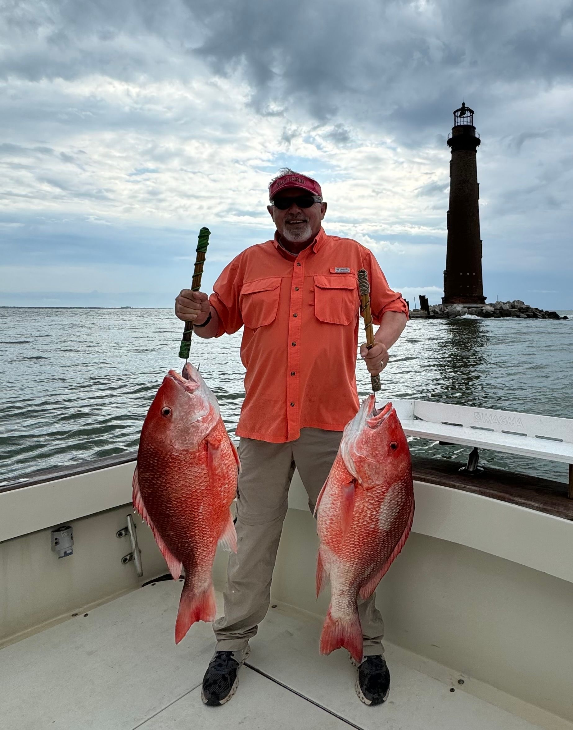 Man on a boat holds two large red fish with a lighthouse behind him under cloudy skies
