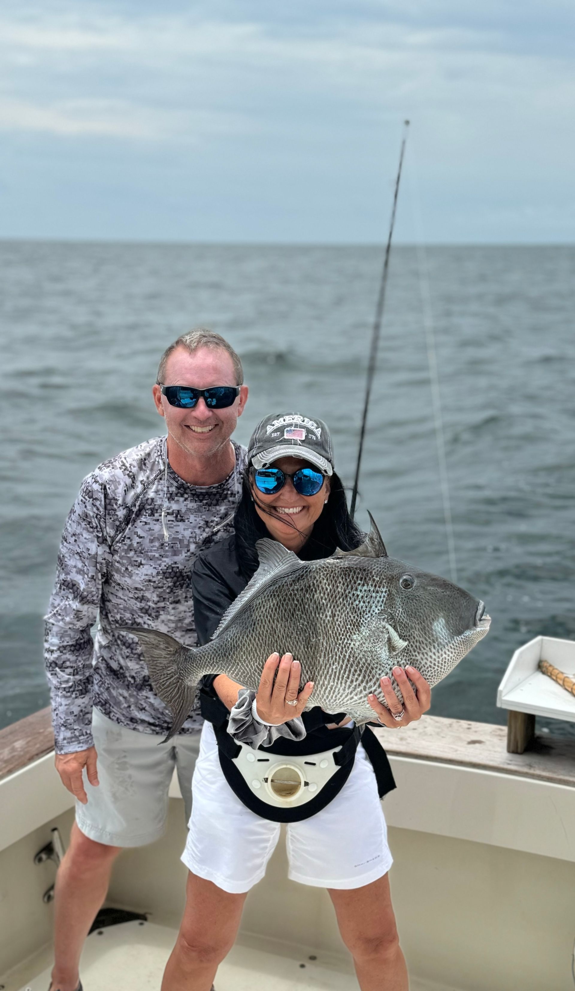 Two people on a boat holding a large fish, with fishing rods and open water behind them.