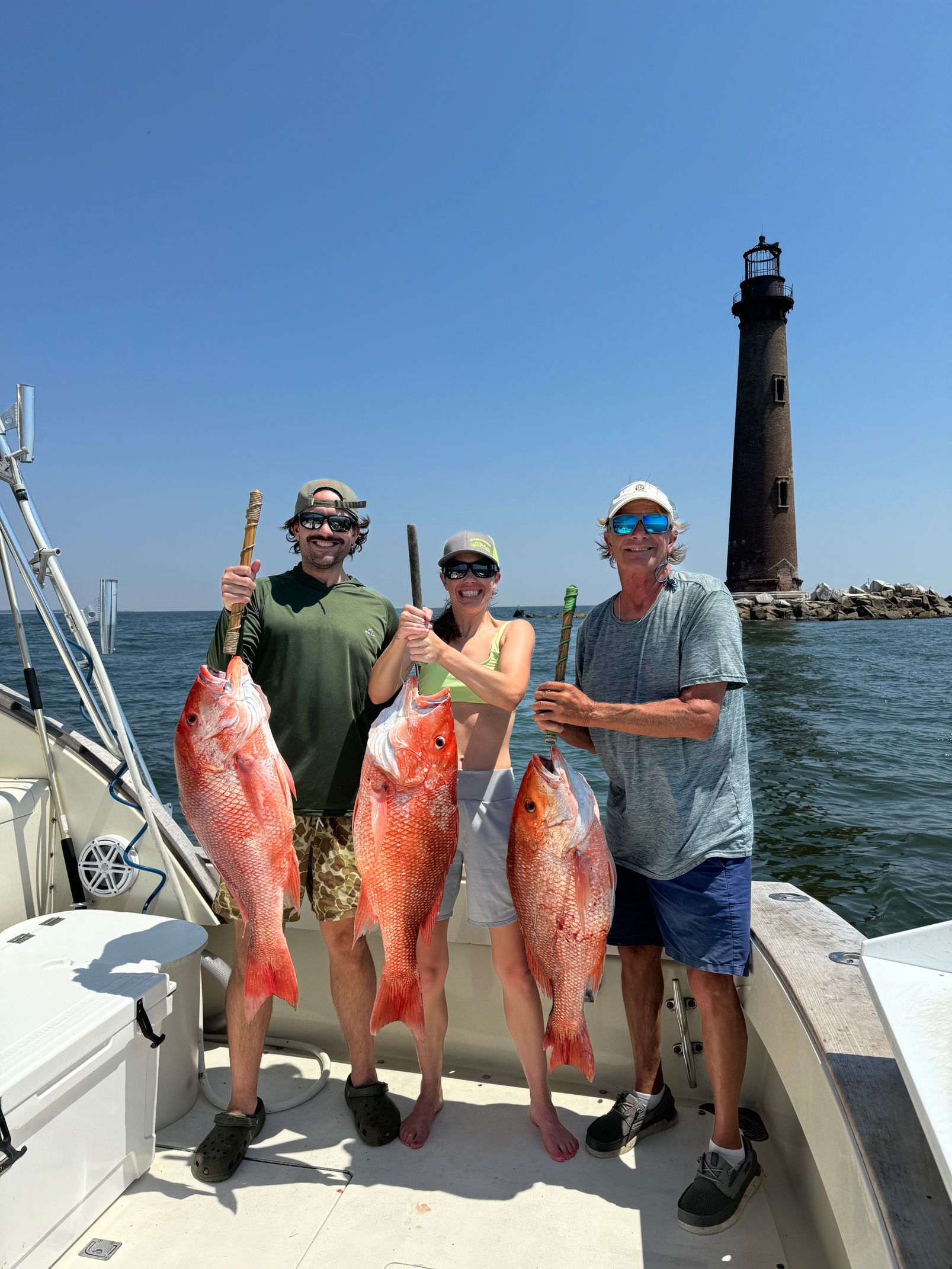 Three people on a boat holding two large red fish with a lighthouse in the background.