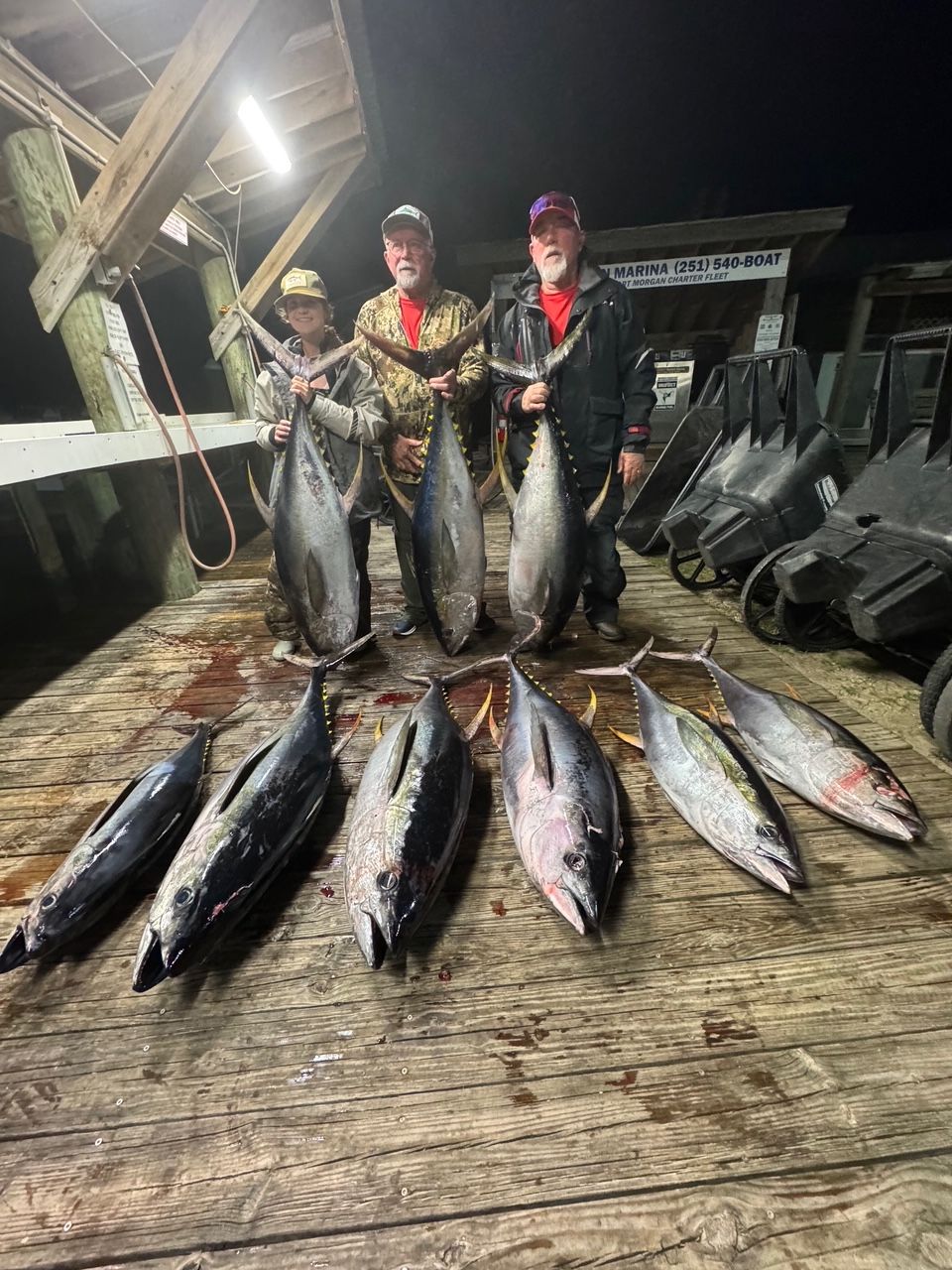 Two anglers holding large fish on a dock at night, with more fish laid out in front of them.