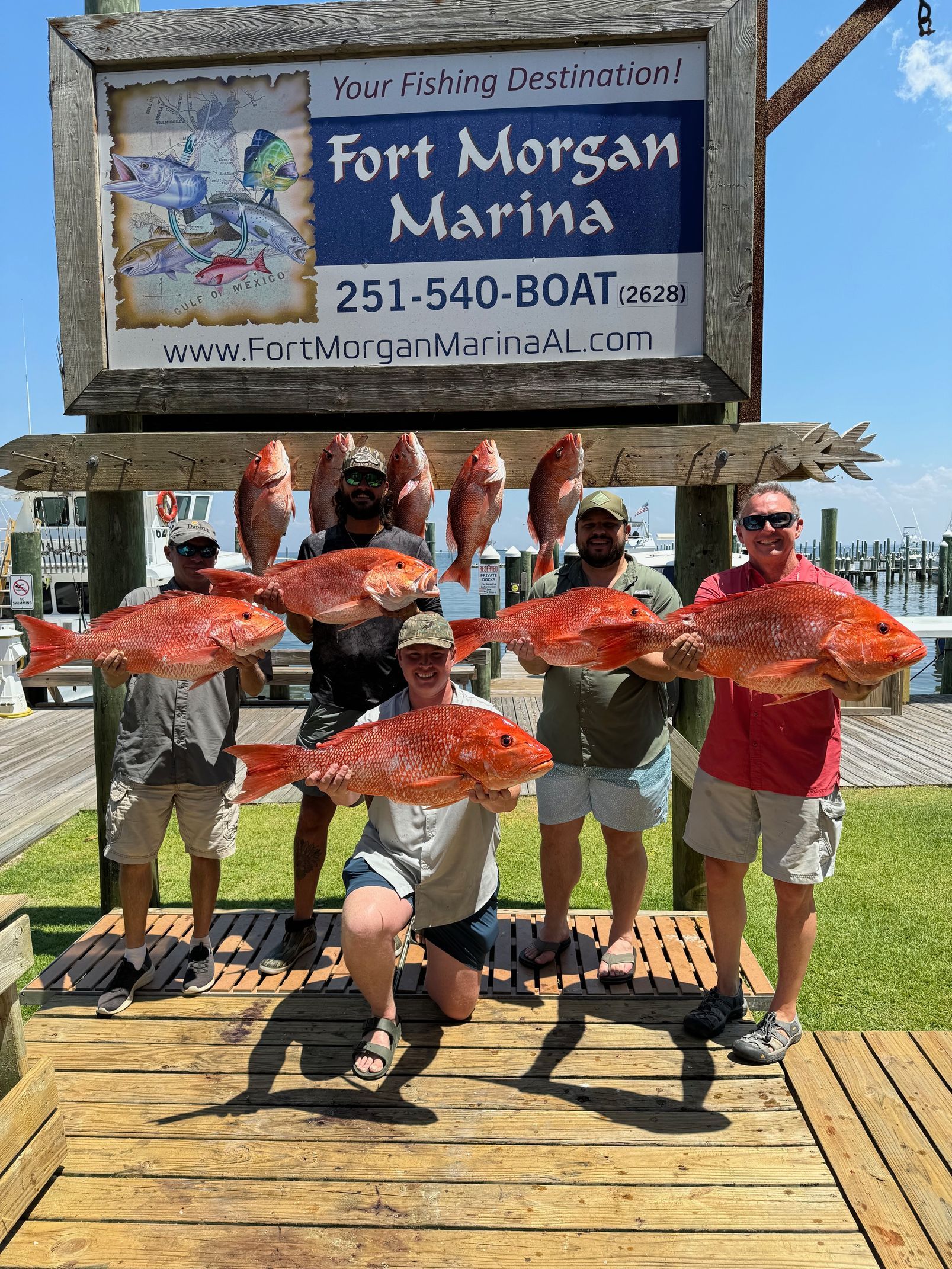 Five people at Fort Morgan Marina holding large red fish under the sign on a sunny dock