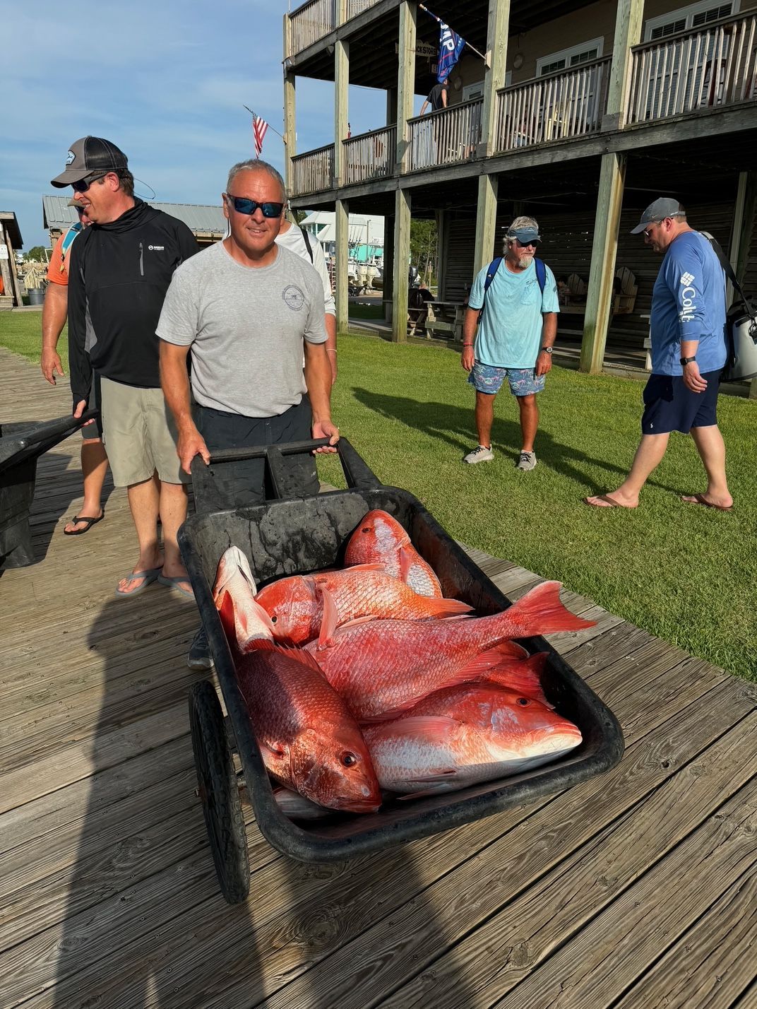 Men hauling large red fish on a tray outdoors by a wooden dock and building.
