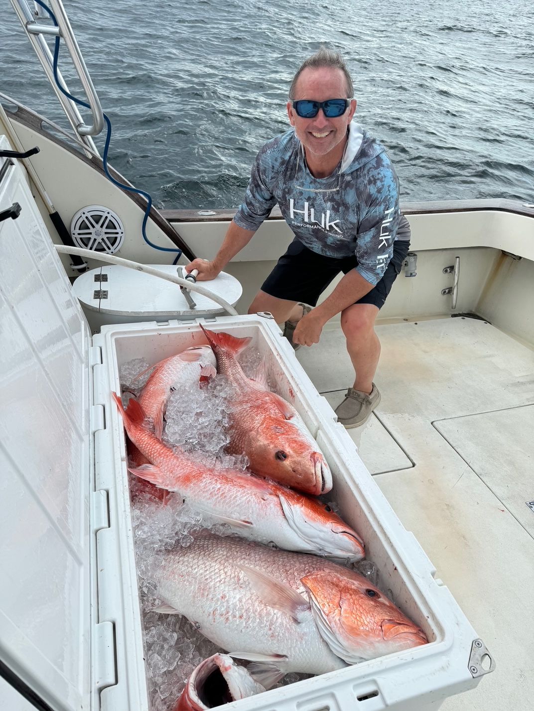 Man aboard a boat smiles beside a cooler full of large fish on the deck with ocean water behind.