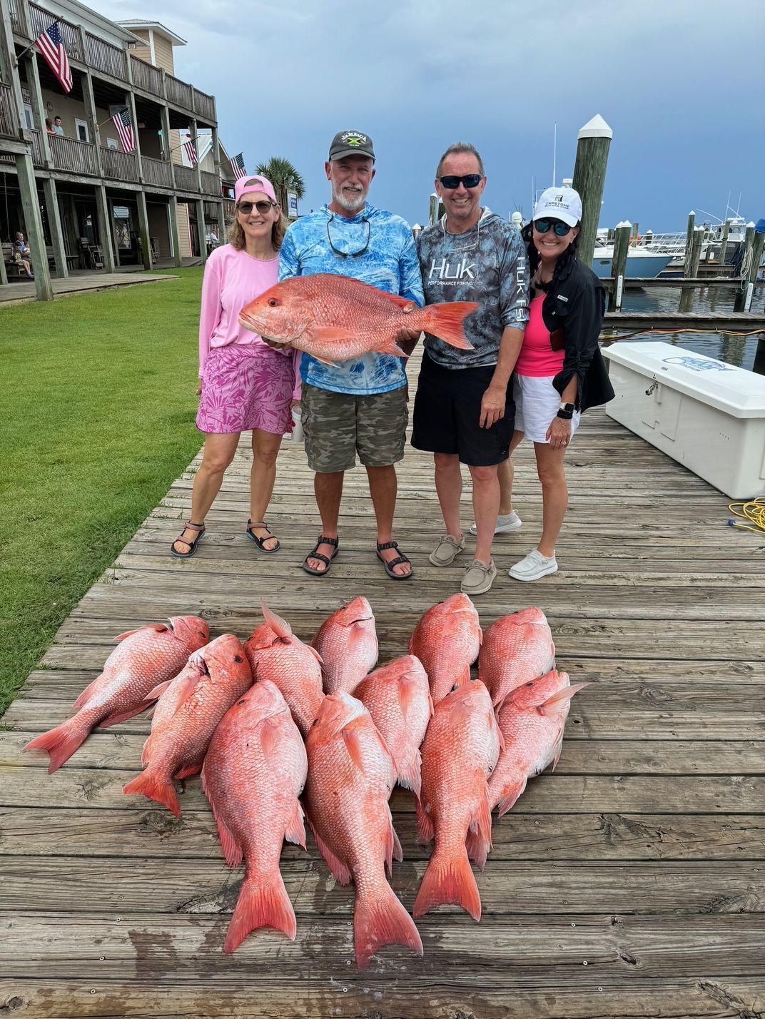 Four people on a dock holding a large red fish, with many red fish laid out on the wooden planks.