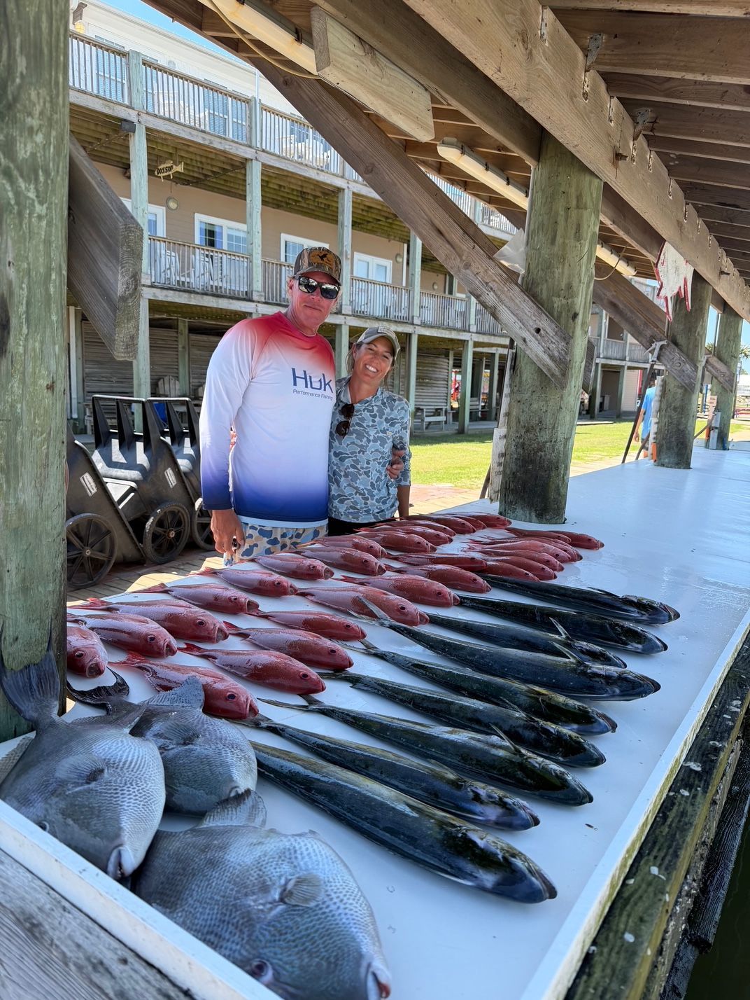 Two people beside a table of freshly caught fish at a dock under wooden beams