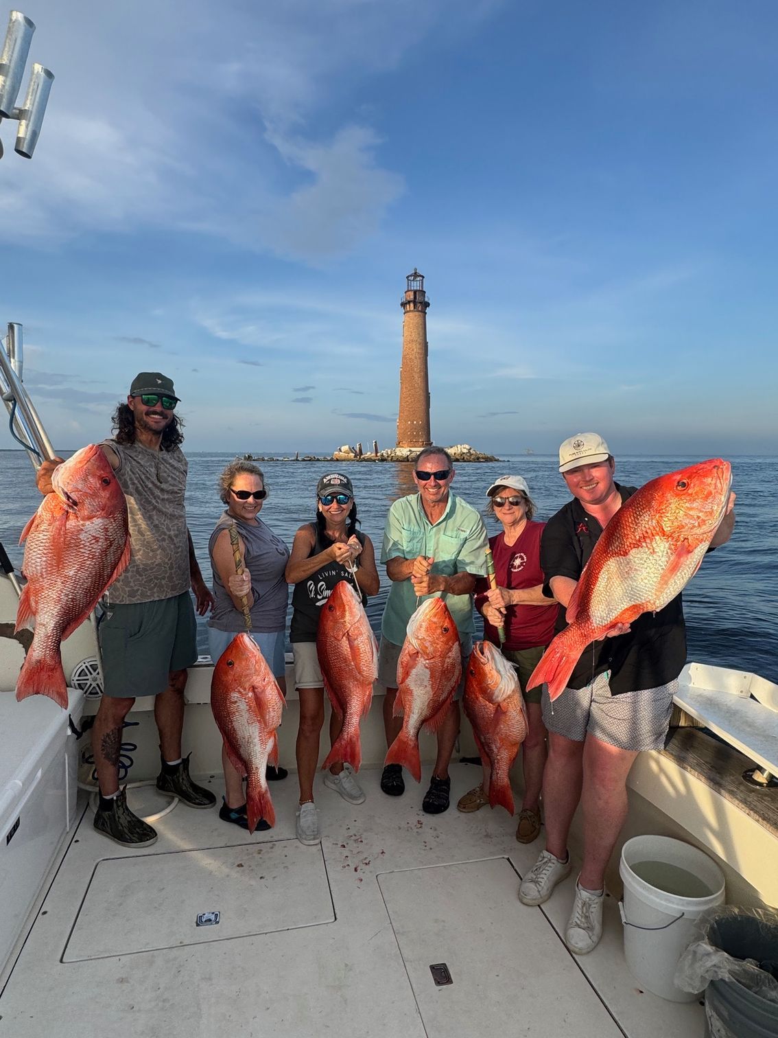 Six people on a boat hold large orange fish with a lighthouse in the background.