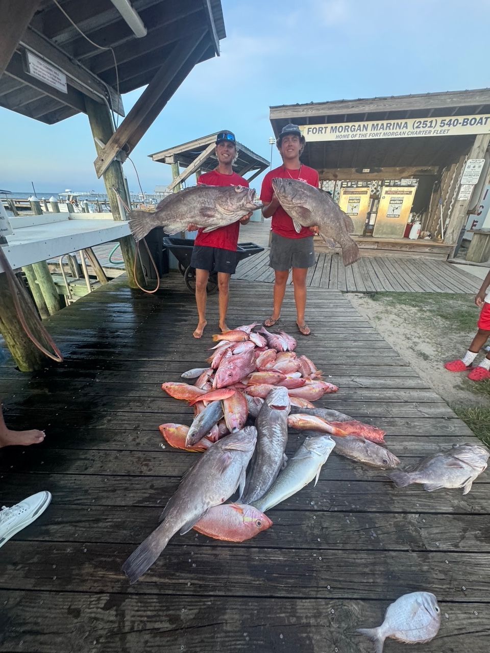 Two people holding fish on a dock beside a large pile of freshly caught fish.