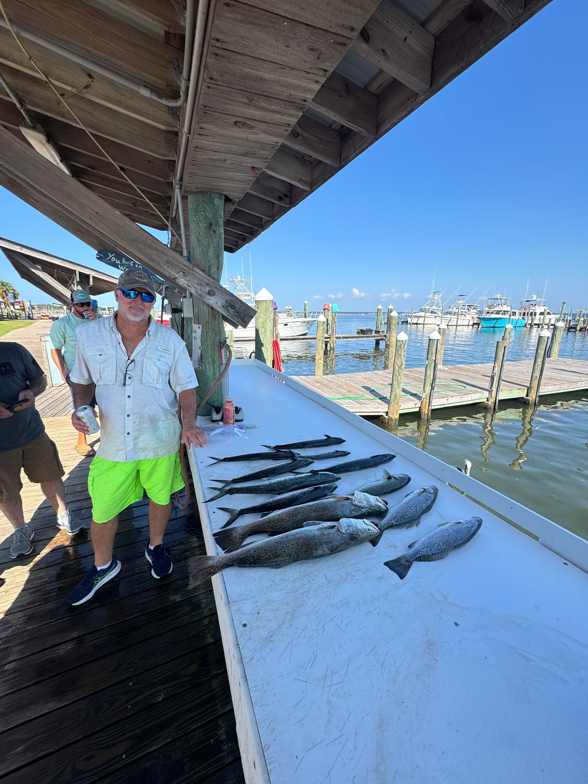 Man standing by a dock with several fish laid out on a white table, boats in the marina behind him.