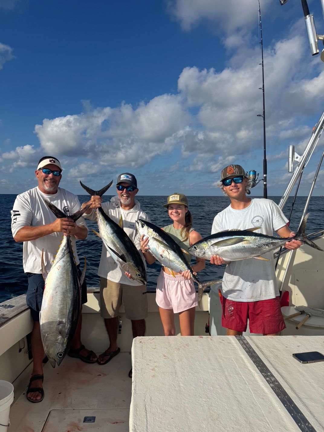 Four anglers on a boat hold up large fish under a bright blue sky with clouds.