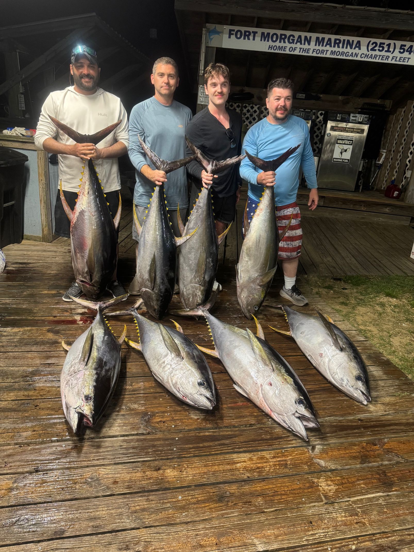 Four people standing with several large fish on a wet dock at night, holding catches for display