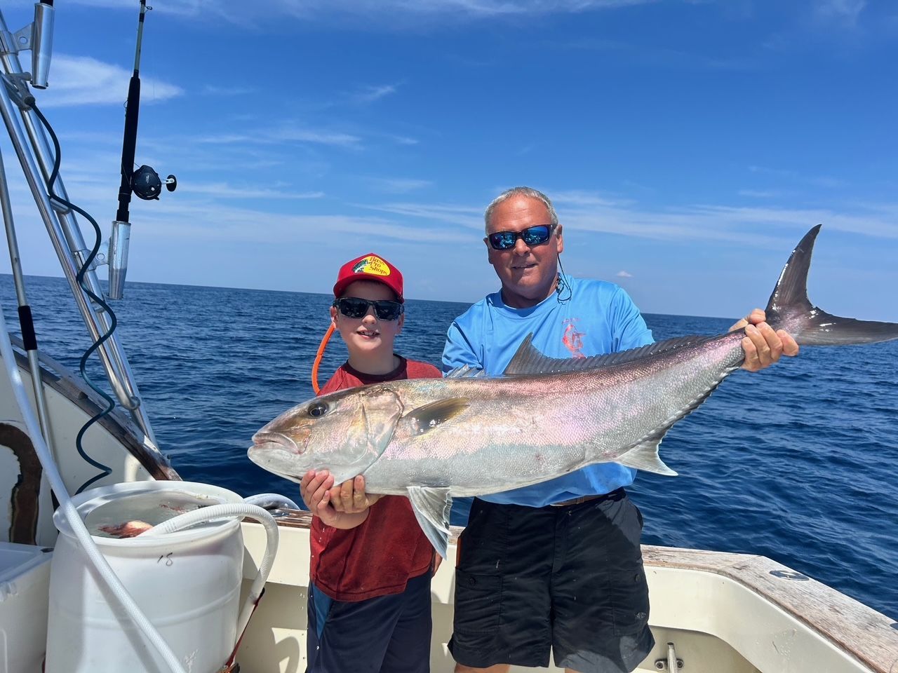 Two people on a boat holding a large silver fish on a sunny blue ocean.