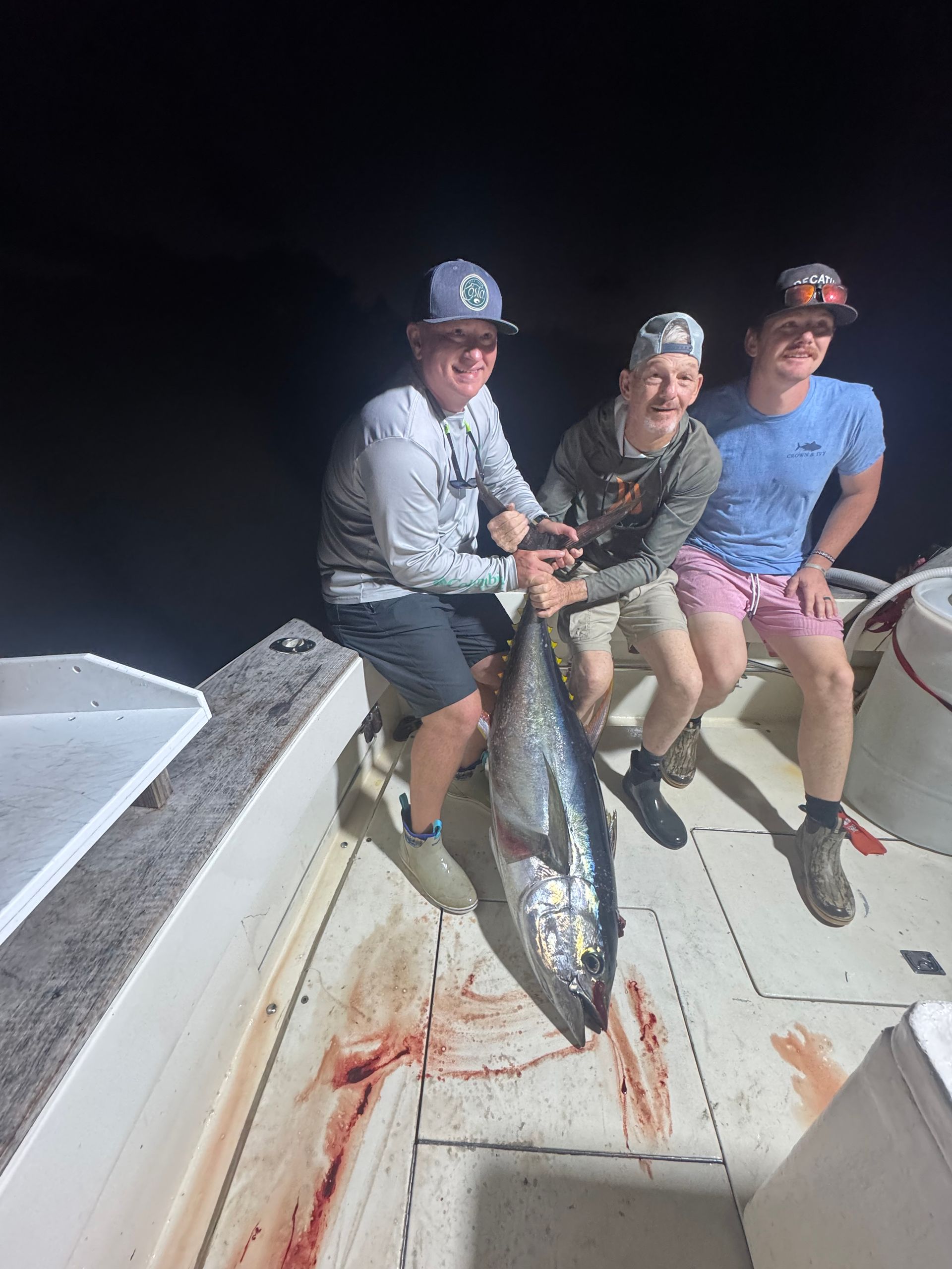 Three people on a boat hold a large fish at night, with blood on the deck.
