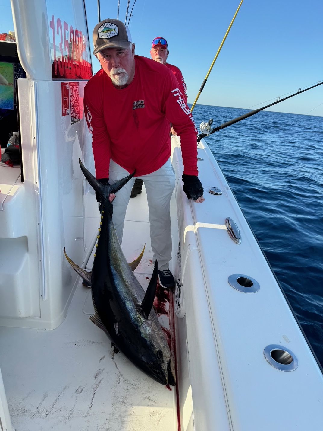 Angler on a boat deck holding a large fish by the tail, with ocean and fishing gear in the background.