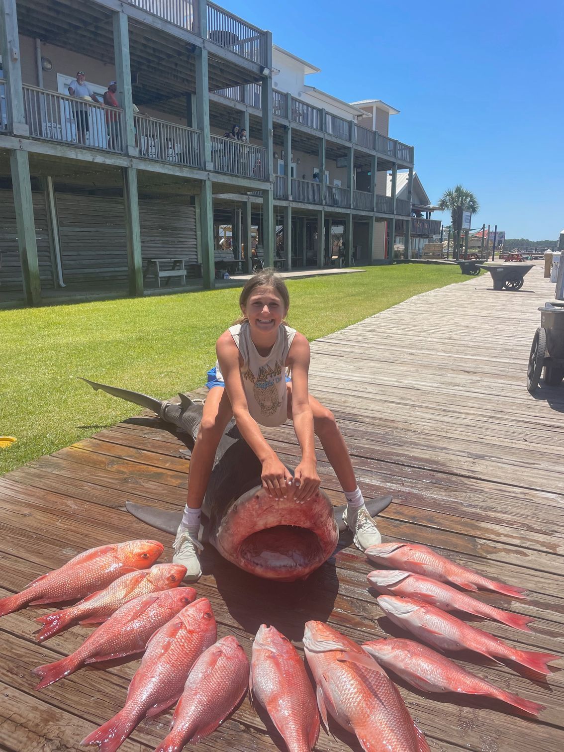 Person kneeling behind a large red fish display on a boardwalk by the water and marina buildings