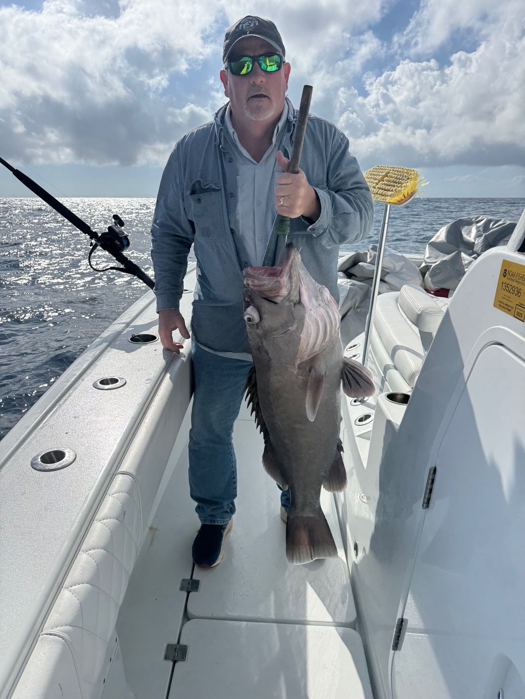 Man standing on a boat deck holding a large fish, with ocean and cloudy sky in the background.