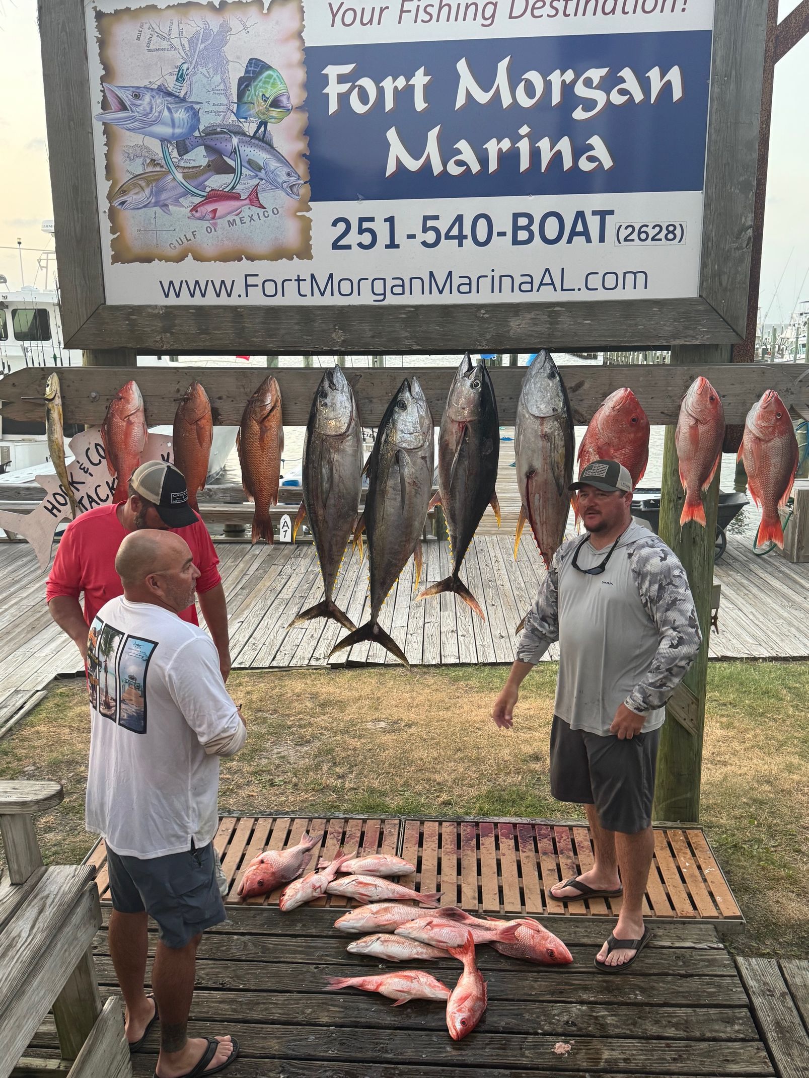 Two people displaying a large catch of fish on a dock beneath a Fort Morgan Marina sign.