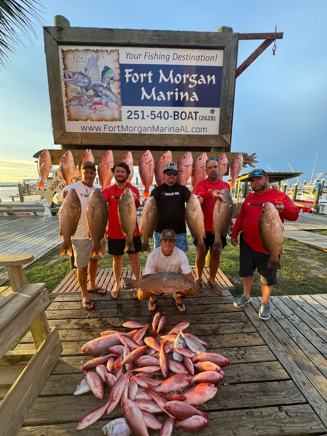 Group posing with a large fish catch on a dock under a Fort Morgan Marina sign at sunset