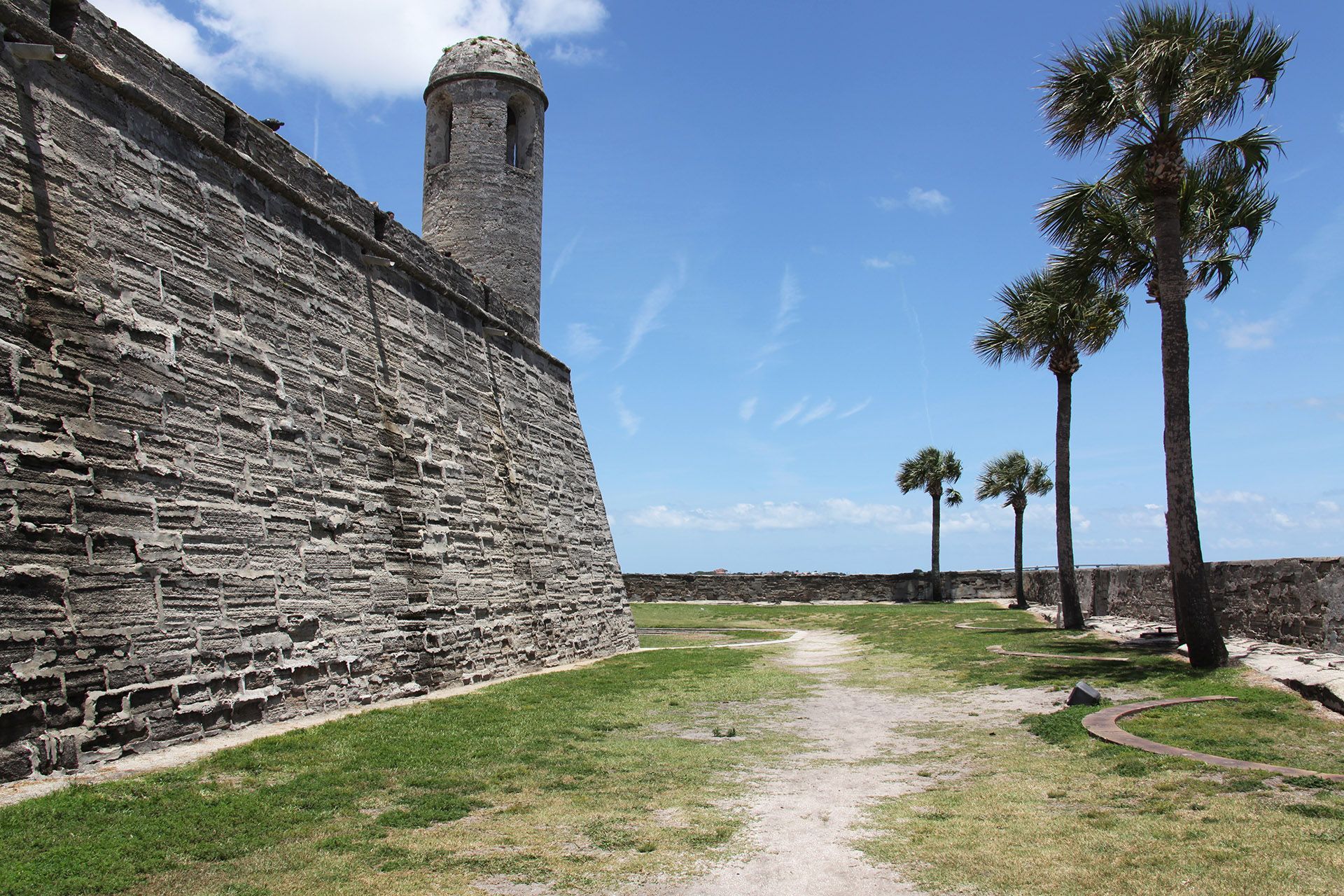 Stone fort with a watchtower, palm trees, and a pathway under a blue sky.