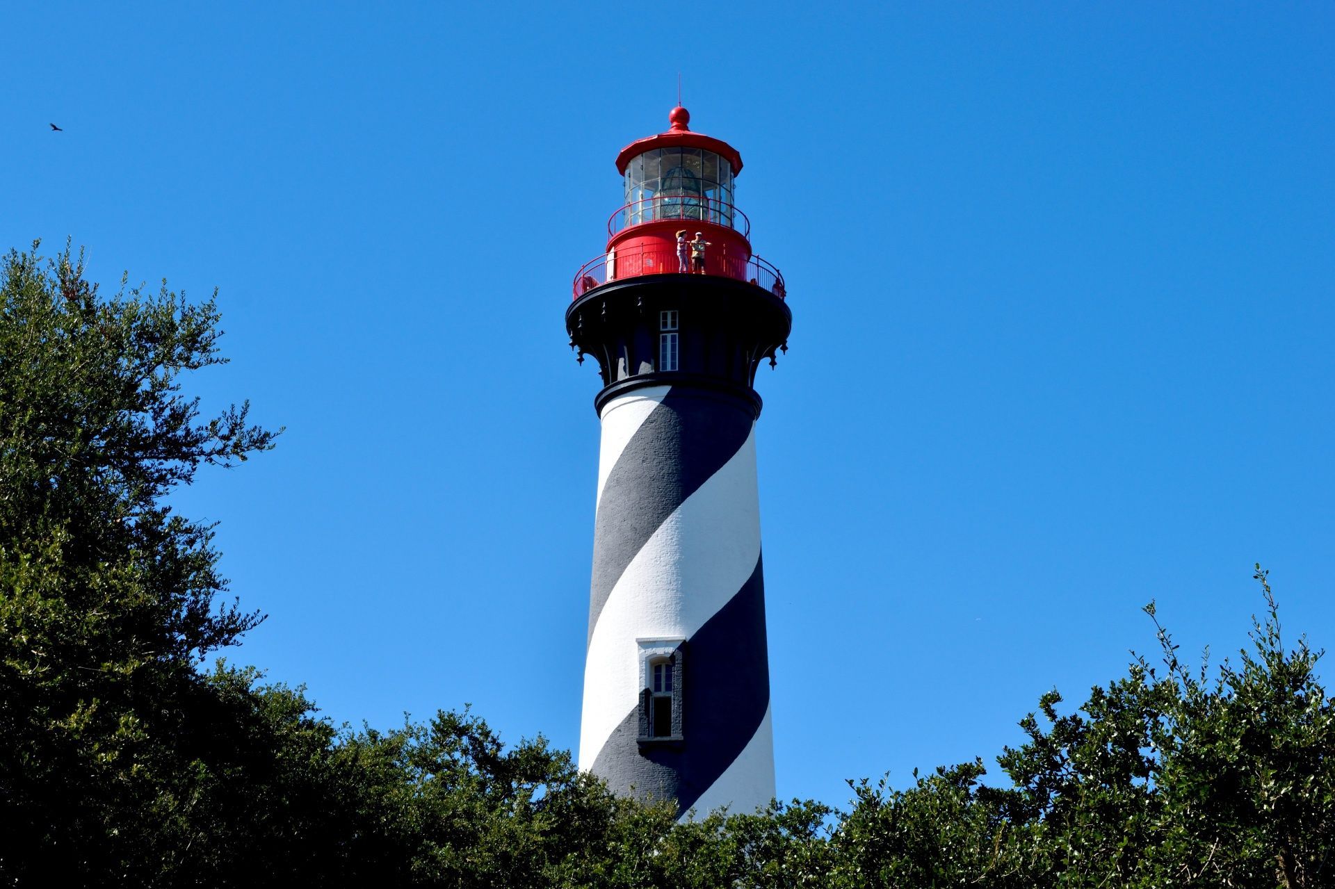 Tall, black and white striped lighthouse with red top, surrounded by green trees, under blue sky.