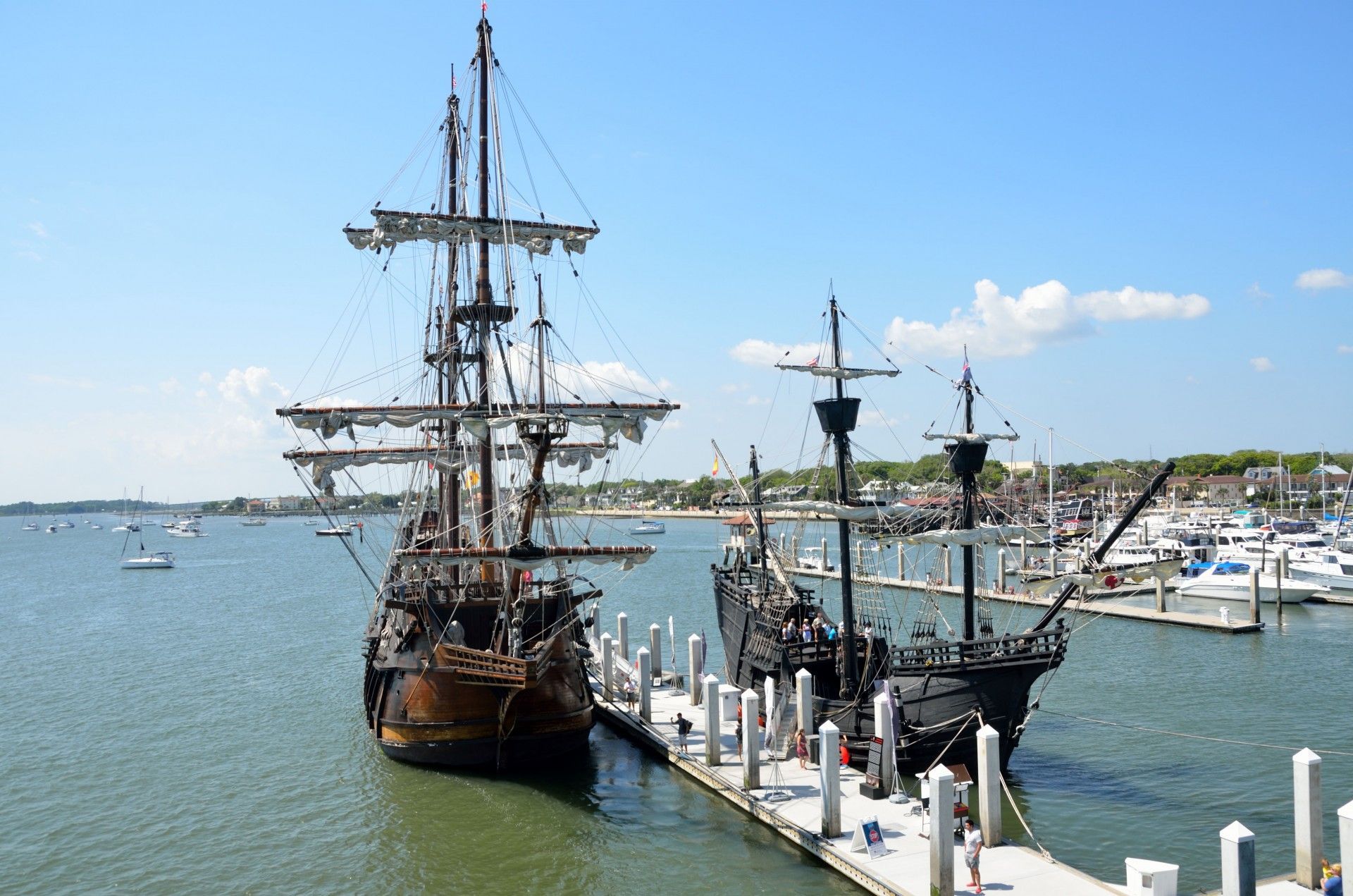 Two tall ships docked side-by-side at a pier in a harbor under a bright blue sky.