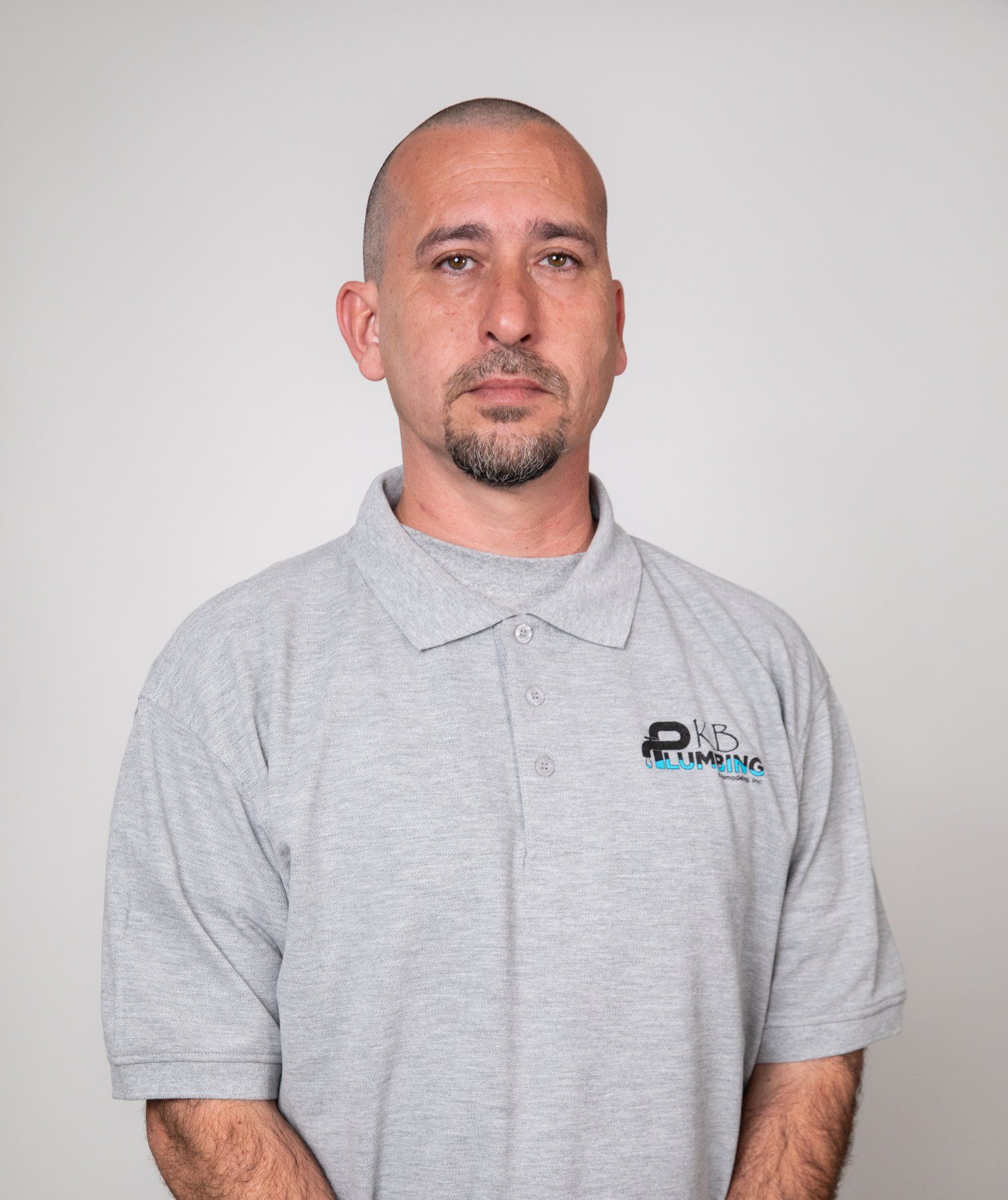 Man with shaved head wearing gray polo shirt, standing in front of a white background.