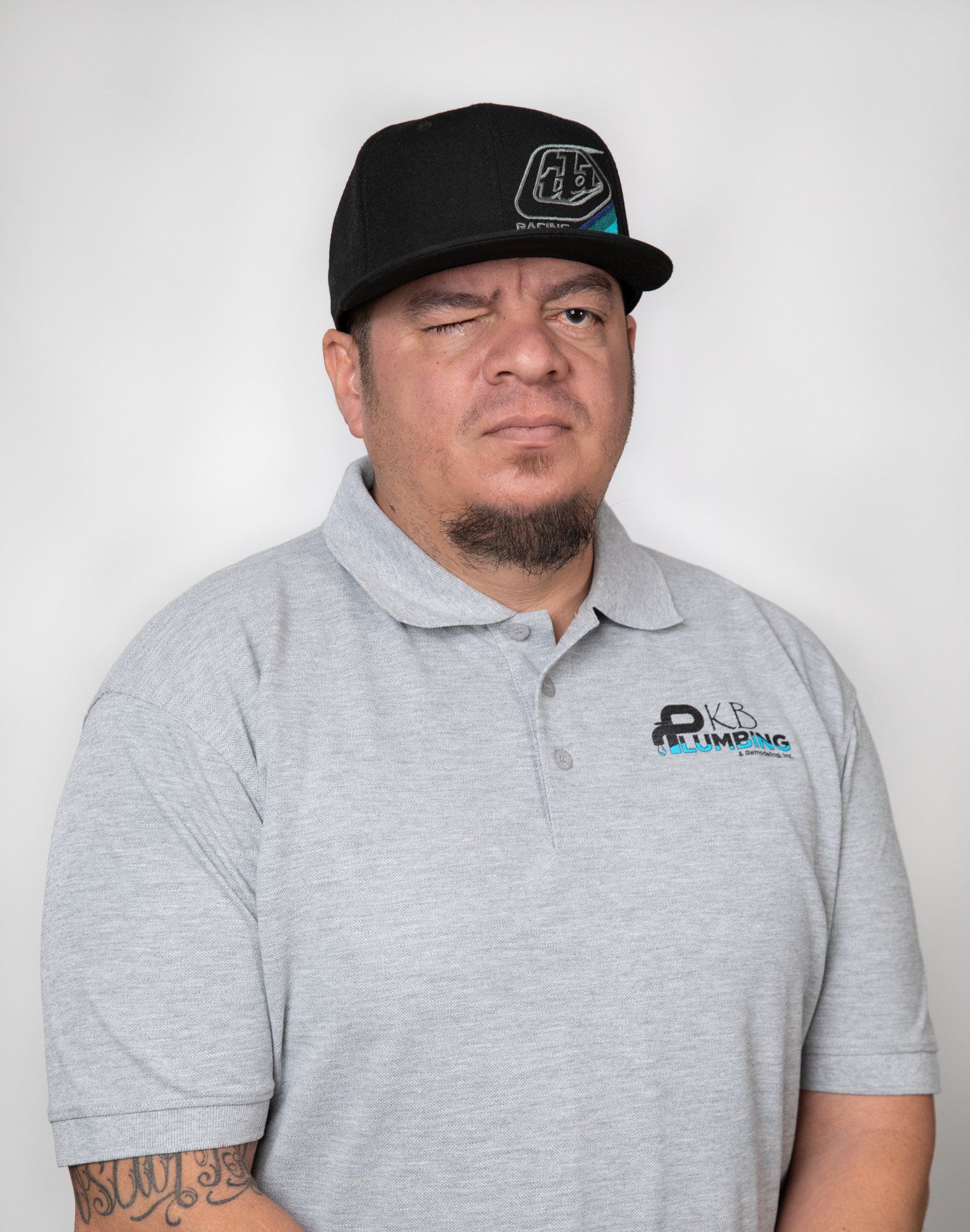 Man in a gray polo shirt and black cap winks, looking at the camera. He has arm tattoos.