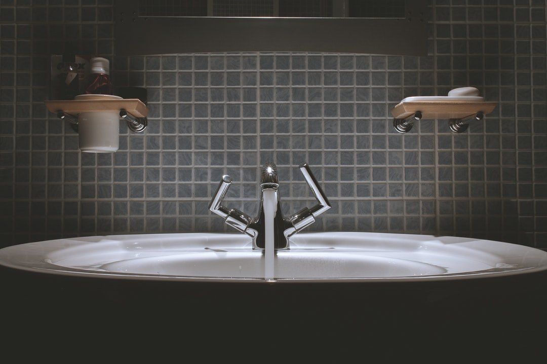 A bathroom sink with a chrome faucet centered, soap dispensers on a tiled wall, dimly lit.
