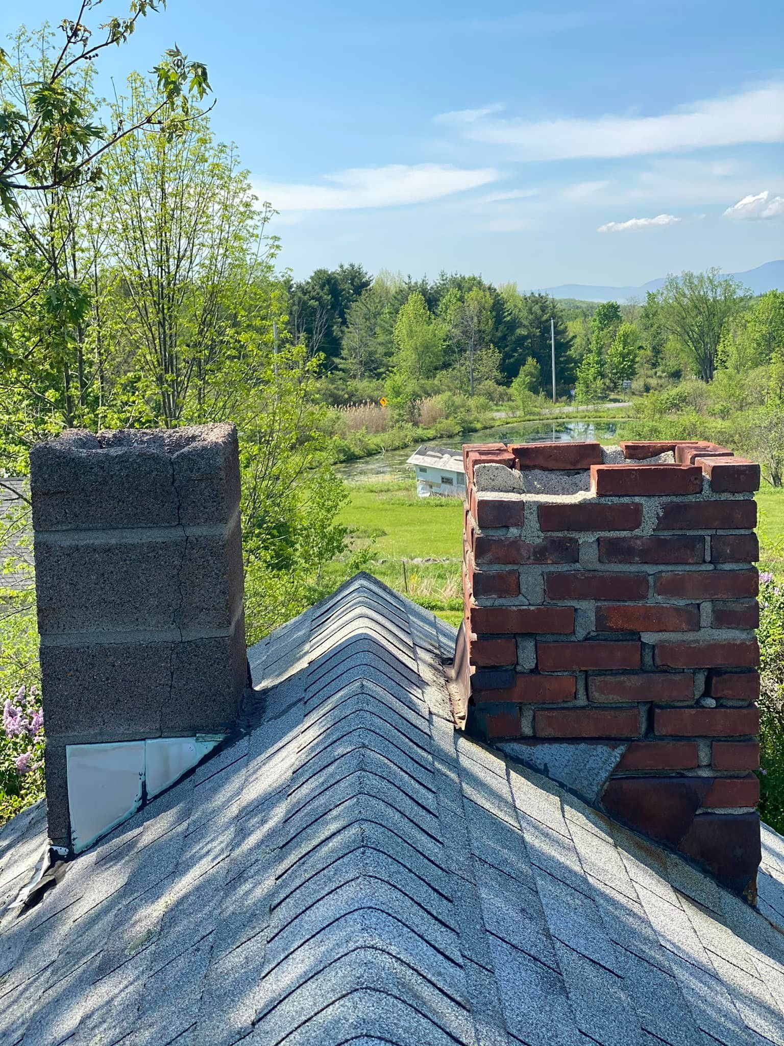 Chimney tops on a roof against a green field and blue sky. One is brick, one is concrete.