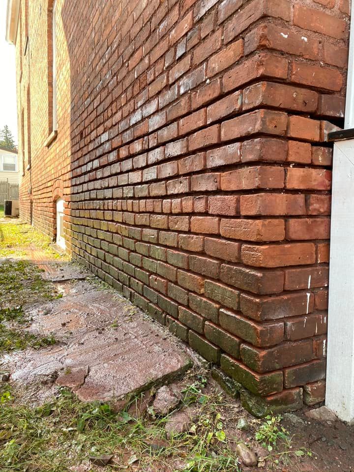Red brick building corner, damp ground with some overgrown vegetation.
