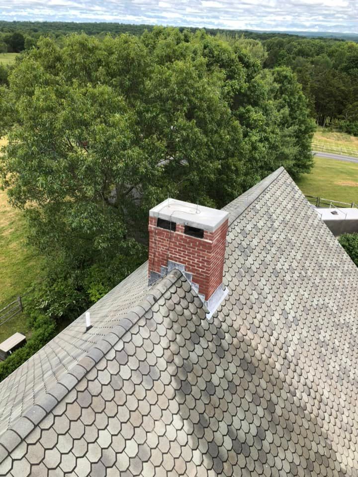 Overhead view of a roof with rounded gray shingles and a brick chimney surrounded by green trees and a cloudy sky.