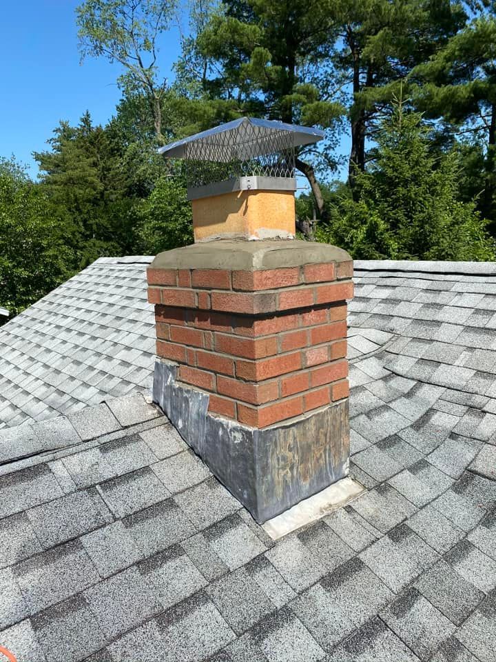 Brick chimney on a gray shingle roof, topped with a metal cap and surrounded by trees under a blue sky.