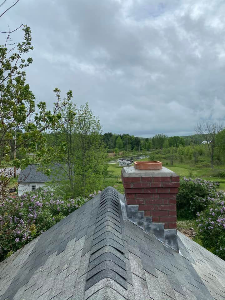 View of a gray shingled roof with a brick chimney, overlooking a green landscape under a cloudy sky.