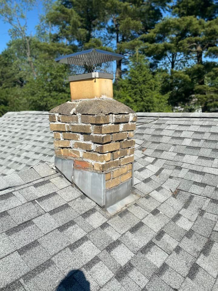 Brick chimney on a gray shingled roof, topped with a metal flue cover.