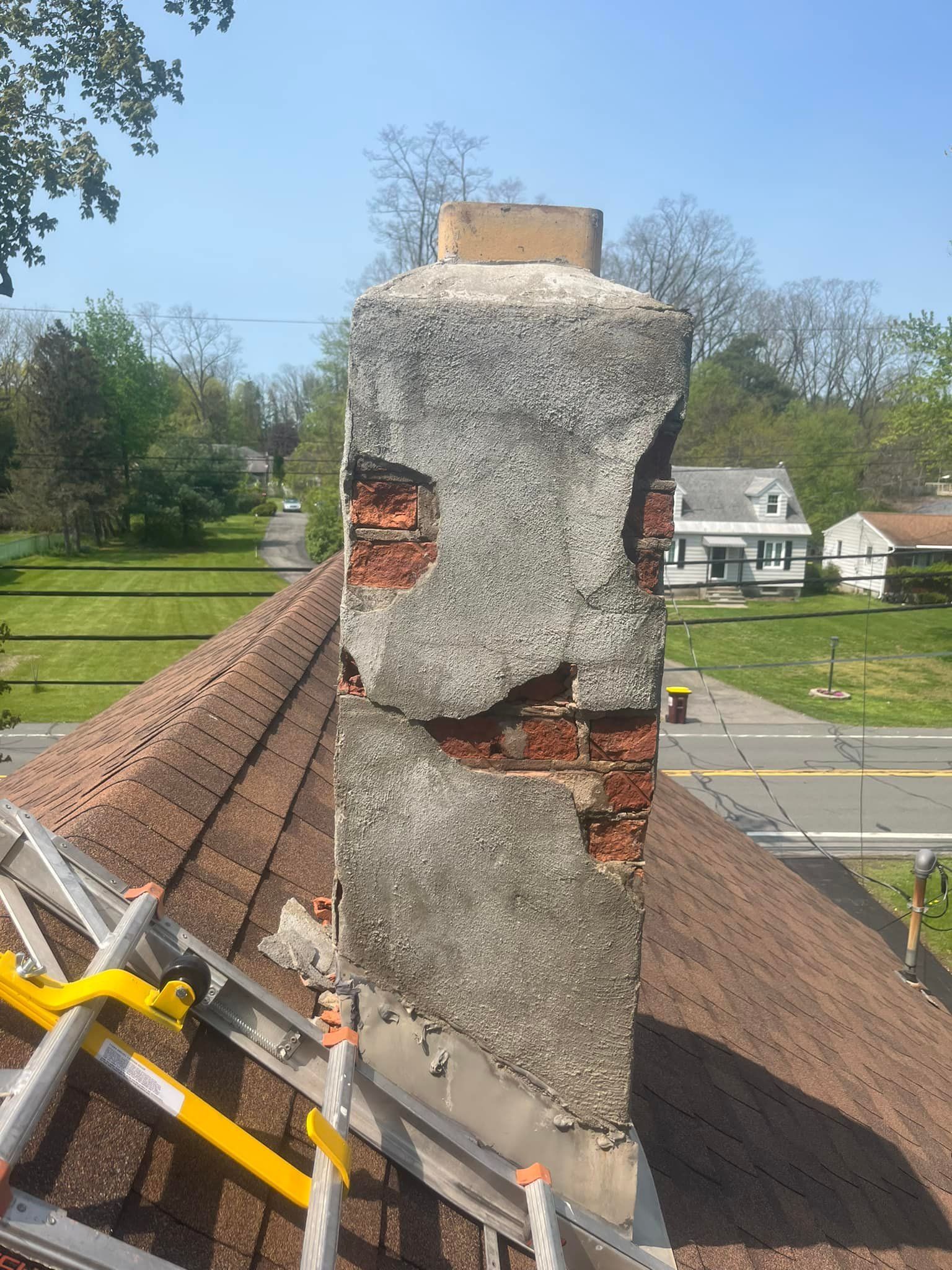 Damaged chimney on a brown shingled roof, showing exposed brick and deteriorating stucco. Houses and trees are in the background.