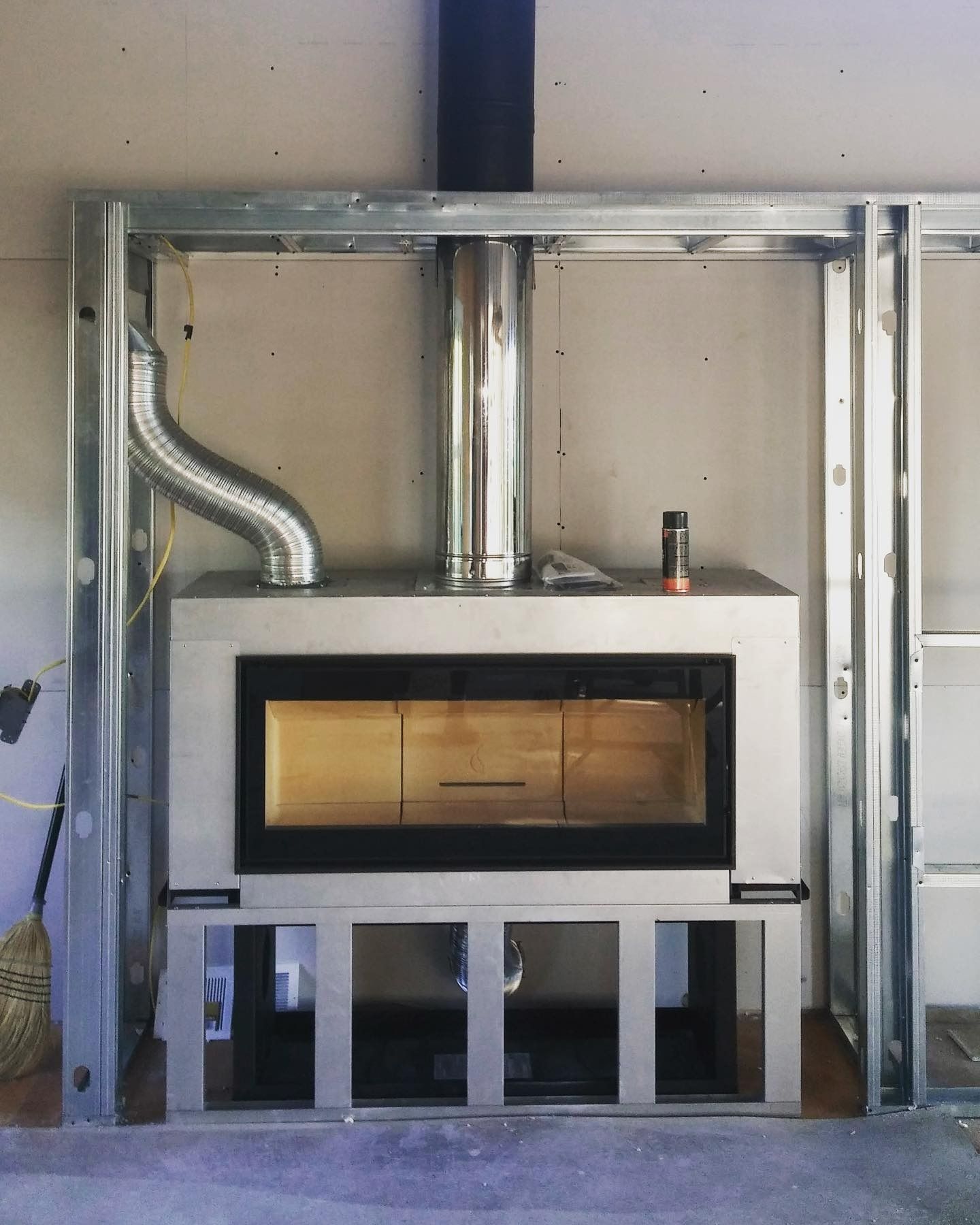 Fireplace inset into metal frame with flue. Silver, rectangular firebox. Chimney and flexible vent pipe visible.