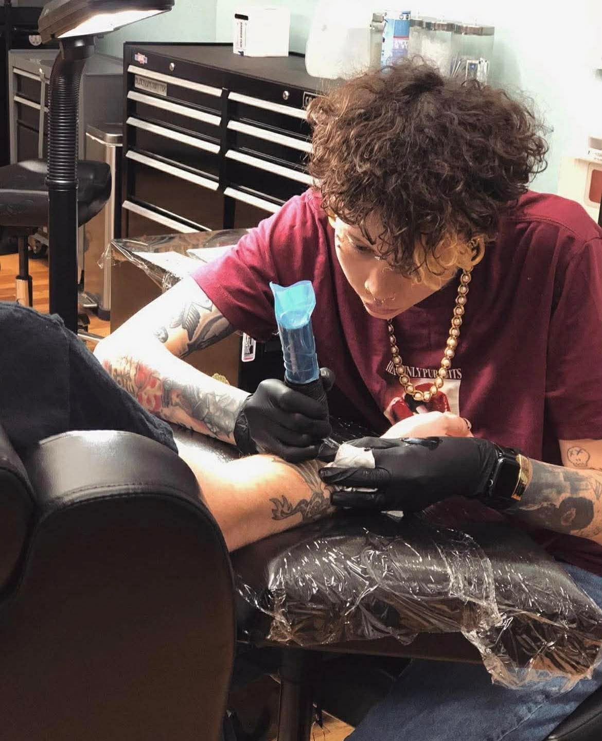 Man with tattoos drinking from a glass, wearing a white t-shirt, hair in a bun. Indoors with dark wall.