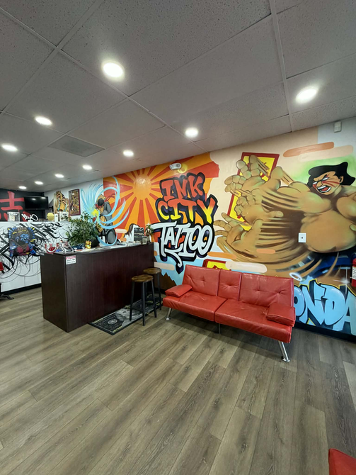 Man with tattoos drinking from a glass, wearing a white t-shirt, hair in a bun. Indoors with dark wall.