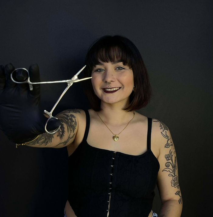 Woman with tattoos holding piercing clamps, smiling at the camera. Black glove, black top, dark background.