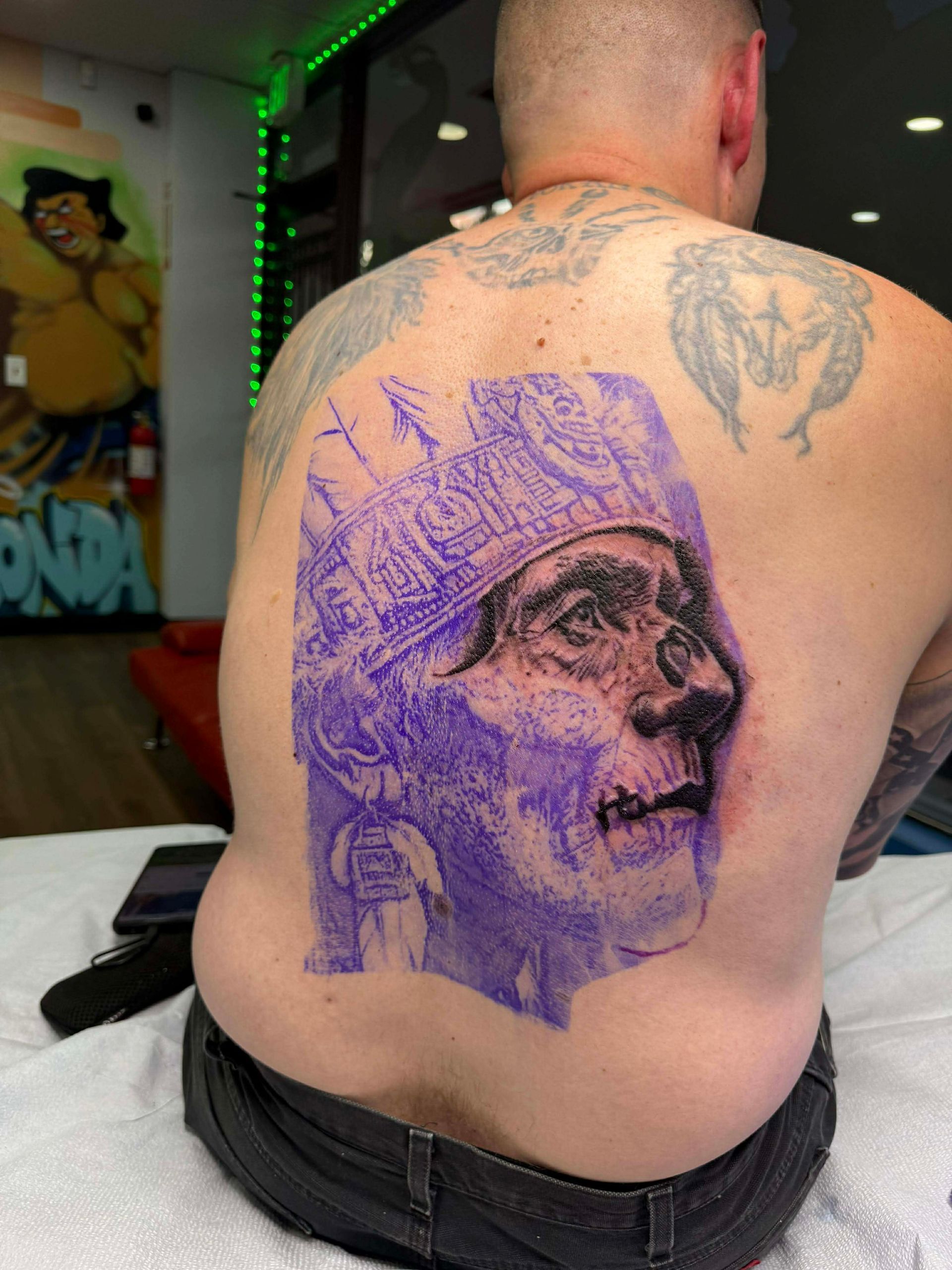 Man's back with a purple transfer of a detailed face tattoo design. Tattoo studio setting.