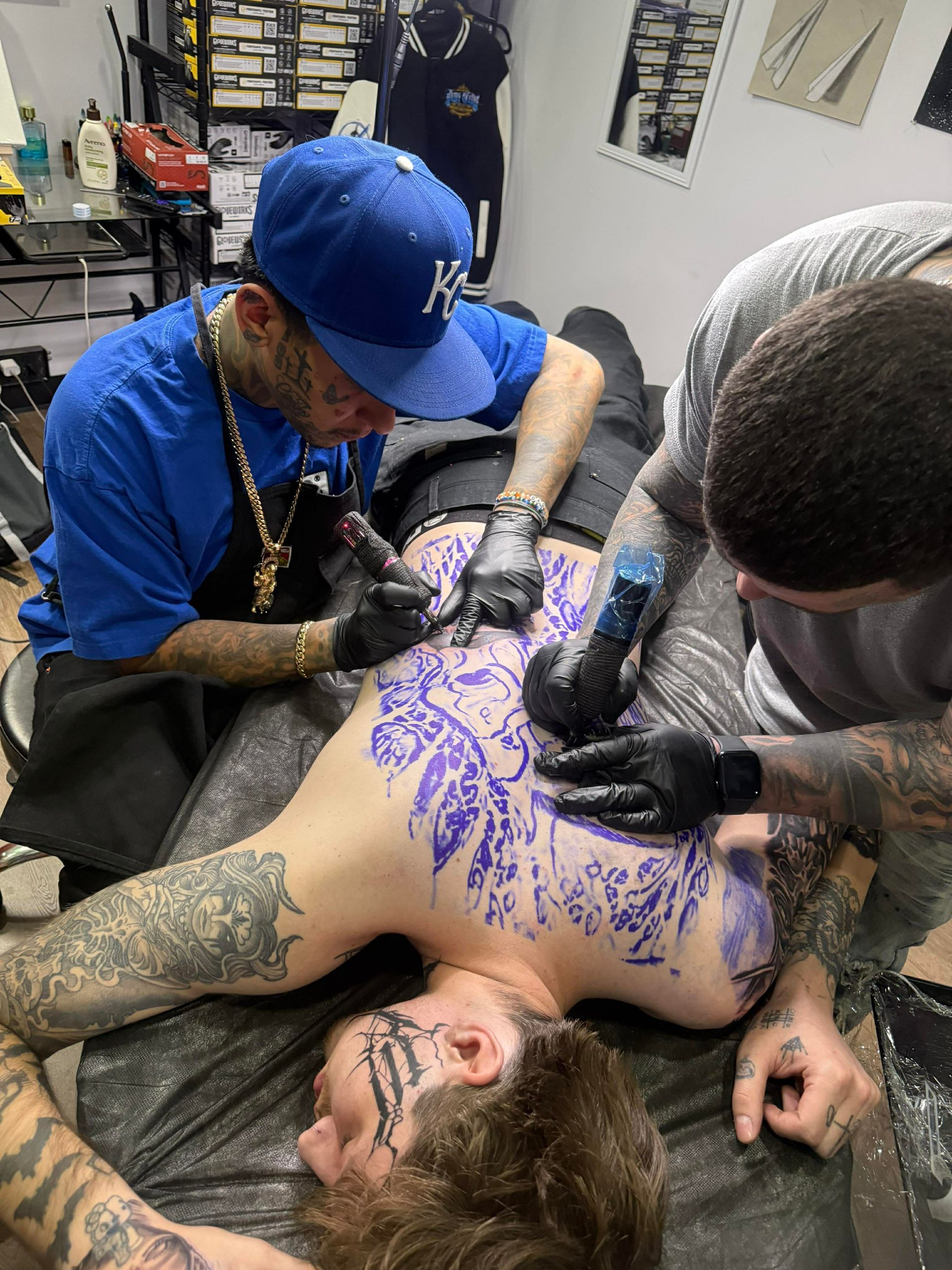 Three people tattooing a person lying down. Tattoo artist with blue hat and two others.