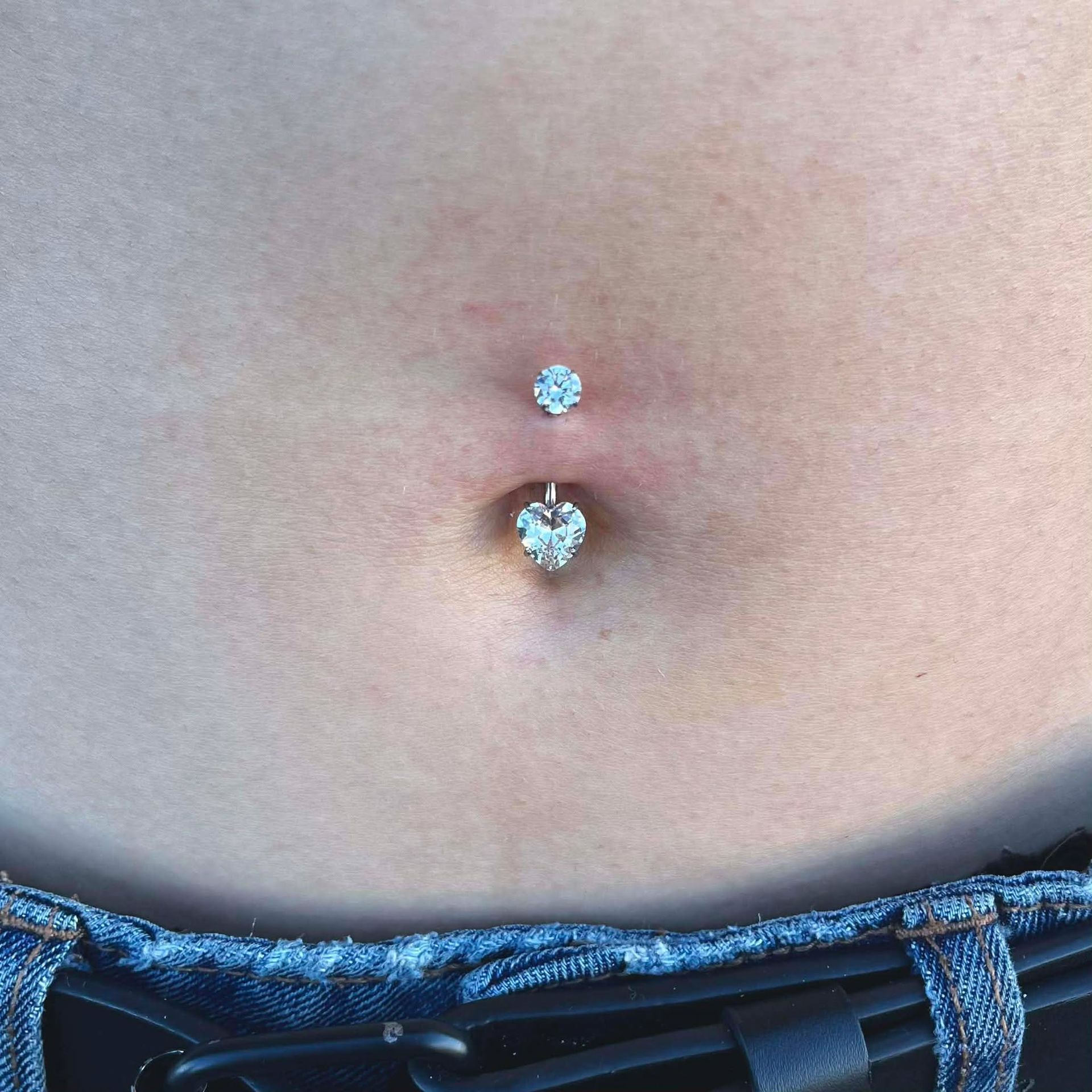 Navel piercing with a sparkling heart-shaped charm and top jewel. Close-up on a person's abdomen.