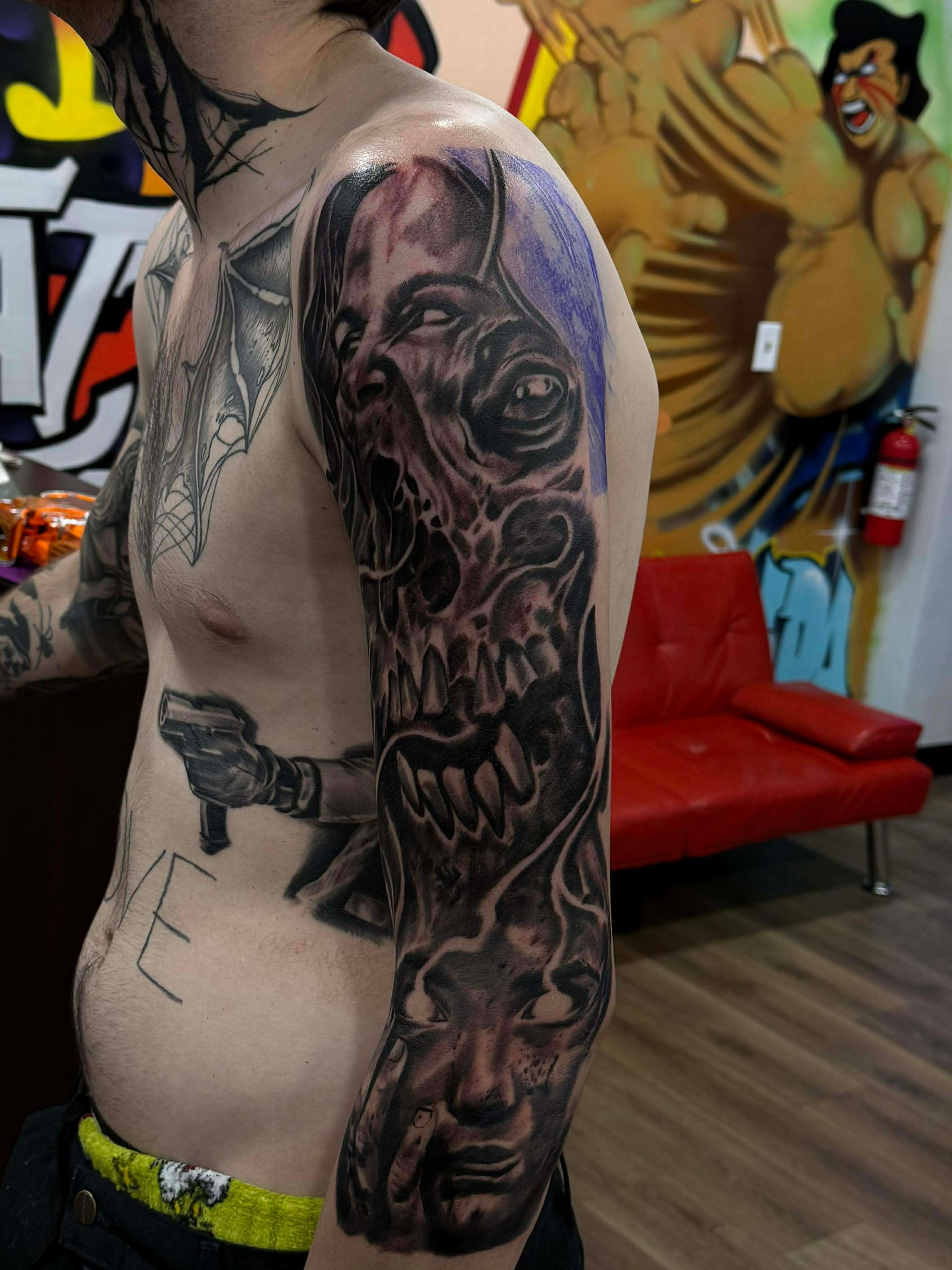 Man with full sleeve tattoo featuring a skull and face imagery in a tattoo parlor.