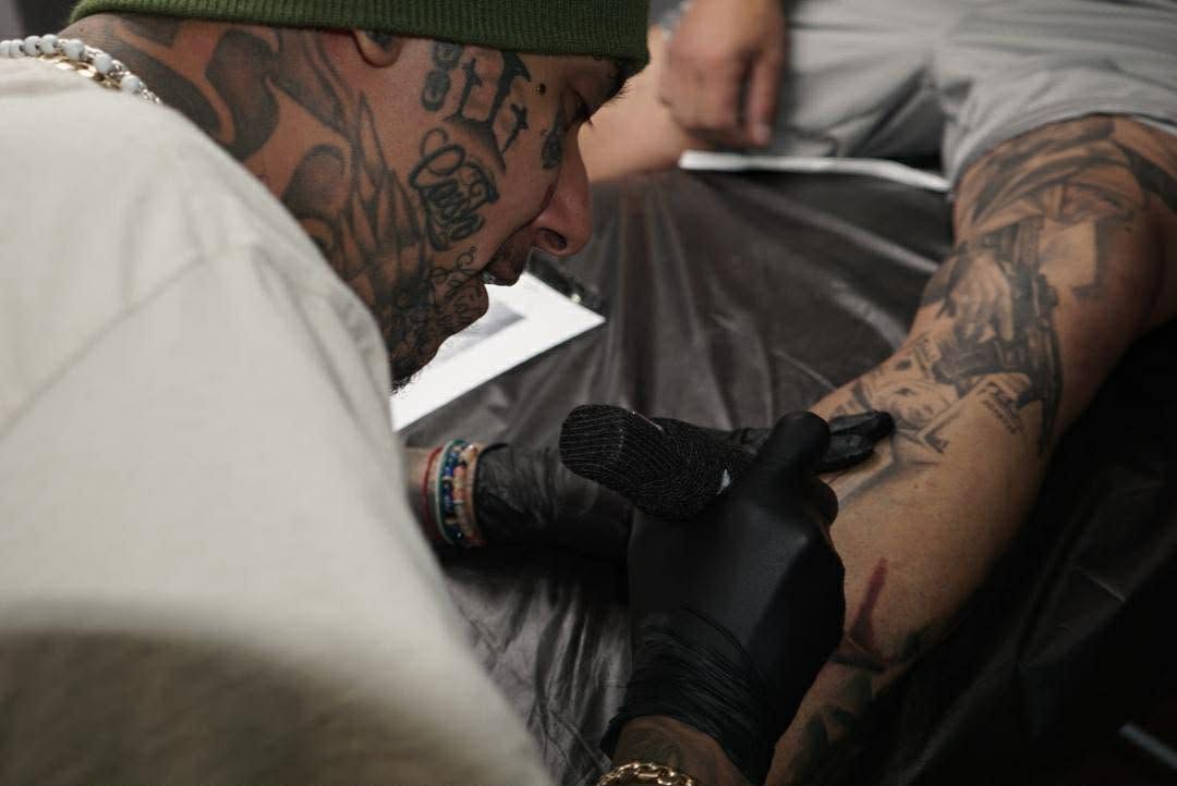 Tattoo artist applying ink to a customer's arm. Artist has face tattoos, wearing gloves, focused.