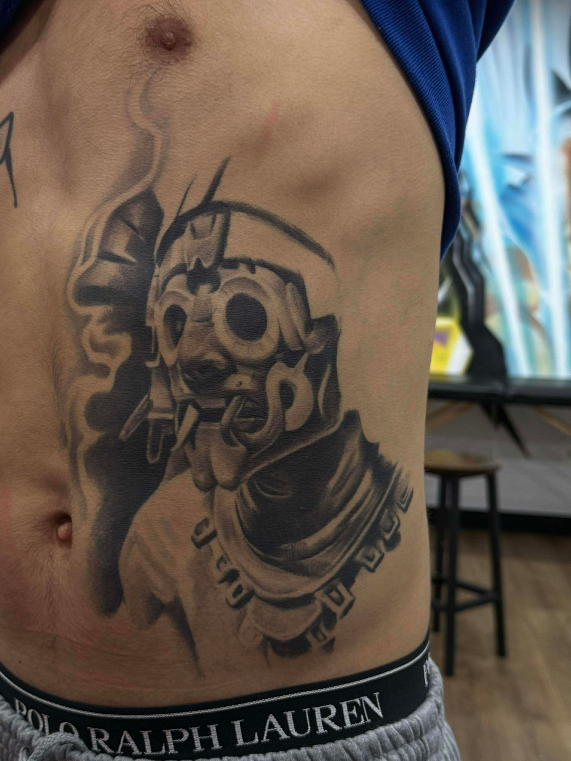 Tattoo of a stylized figure with a mask, on a person's side, in grayscale. Smoke surrounds the figure.