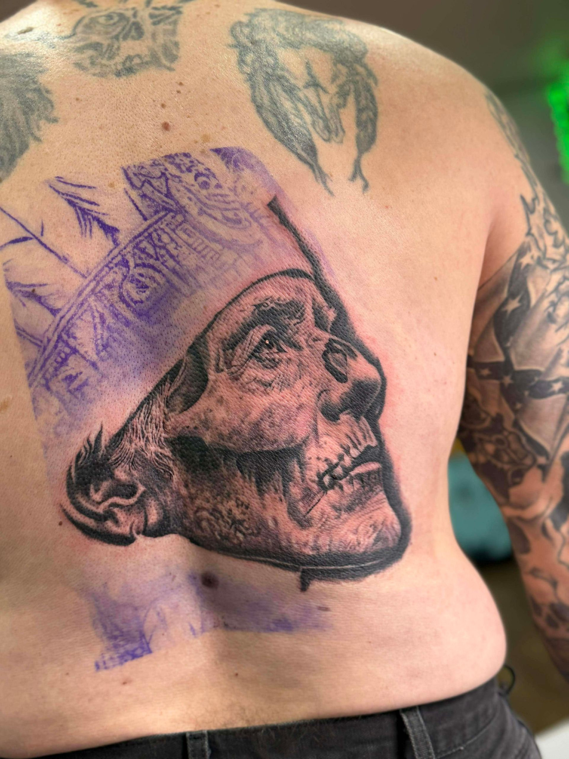 Back tattoo of a skull wearing a hat. Detailed black and white shading, with purple stencil outlines.