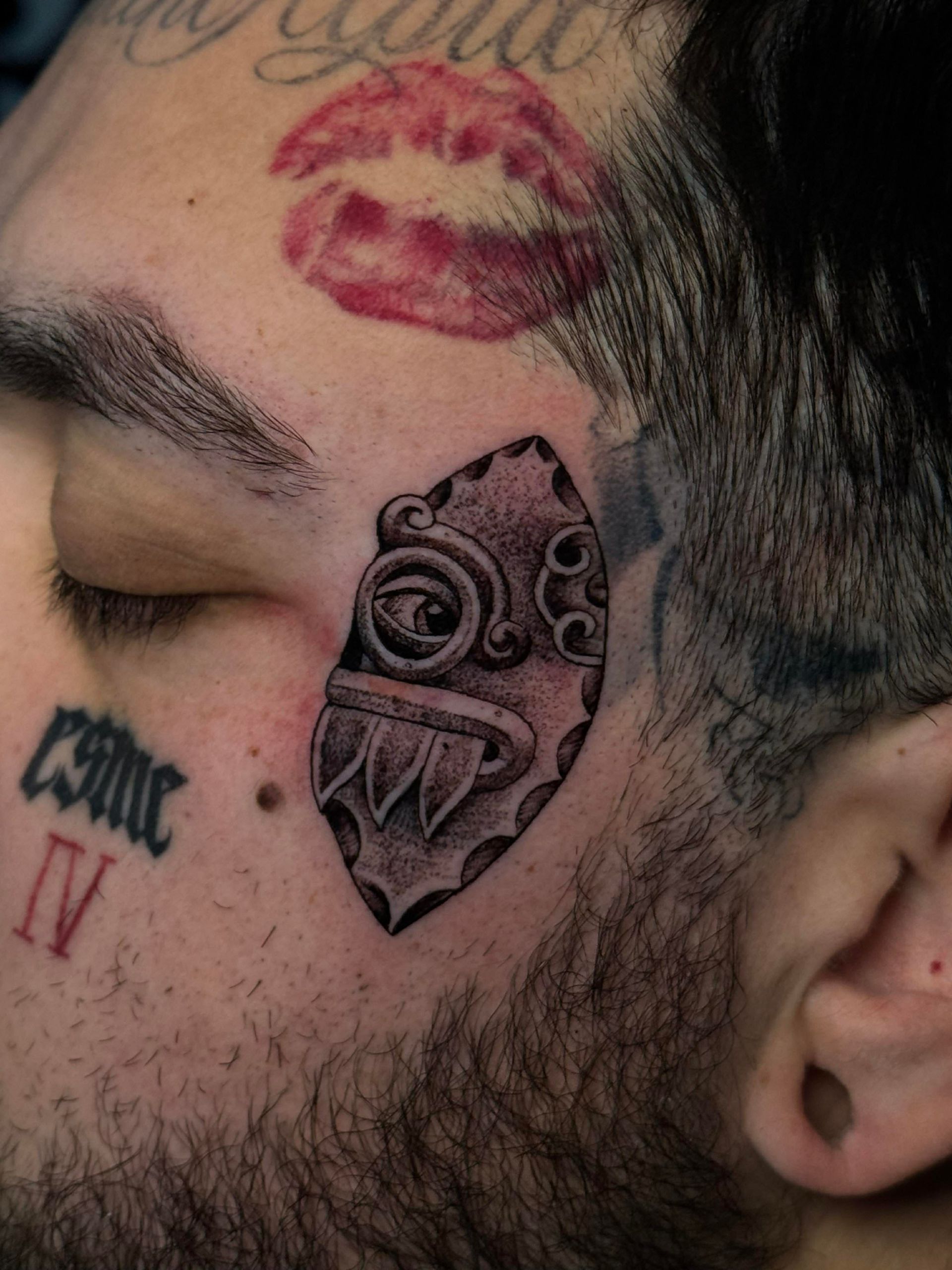Close-up of a person's face with tattoos: a mask, lipstick print, and lettering.