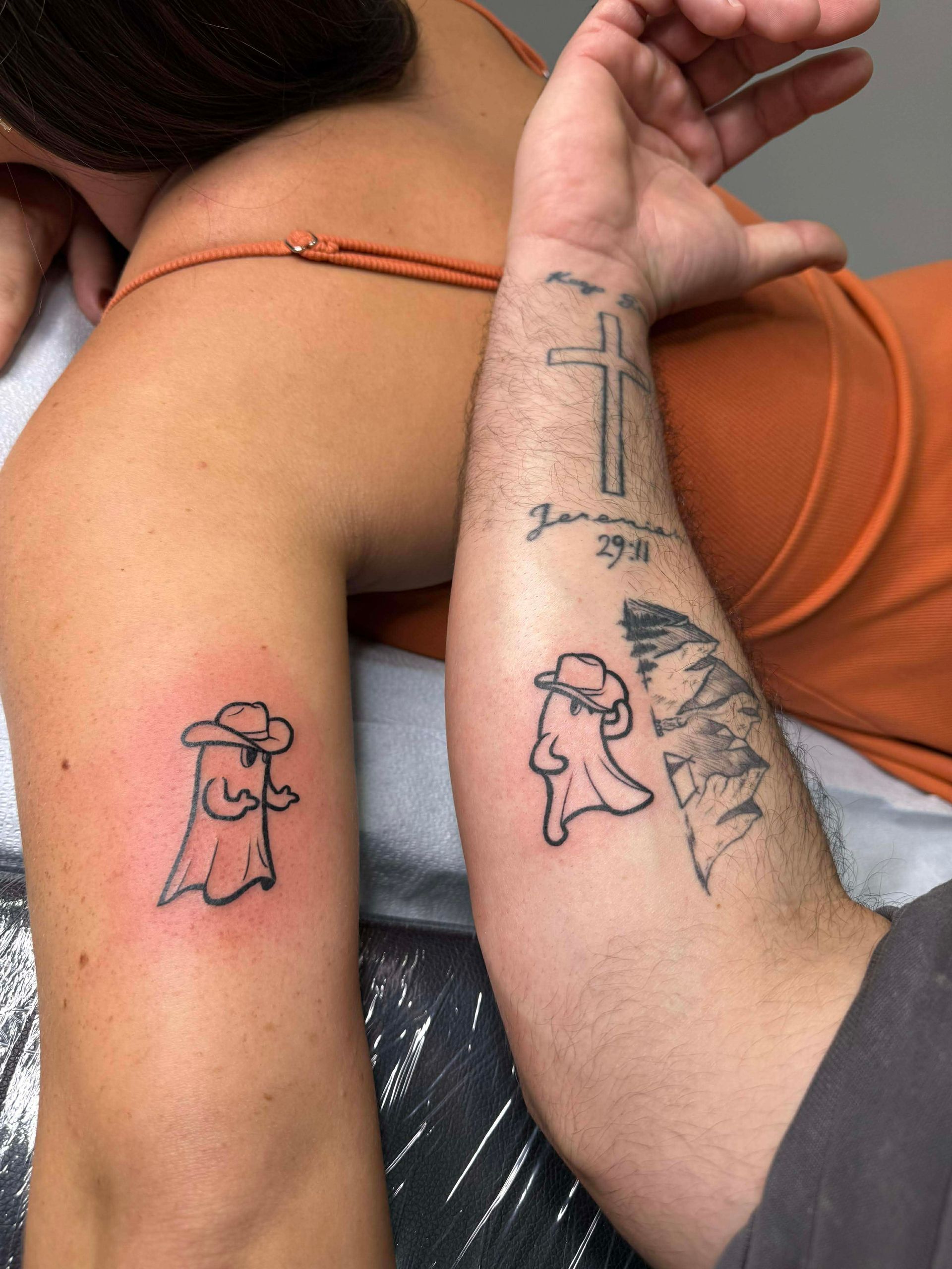 Two arms side-by-side, each with matching cartoon ghost tattoos wearing hats. Person in orange shirt.