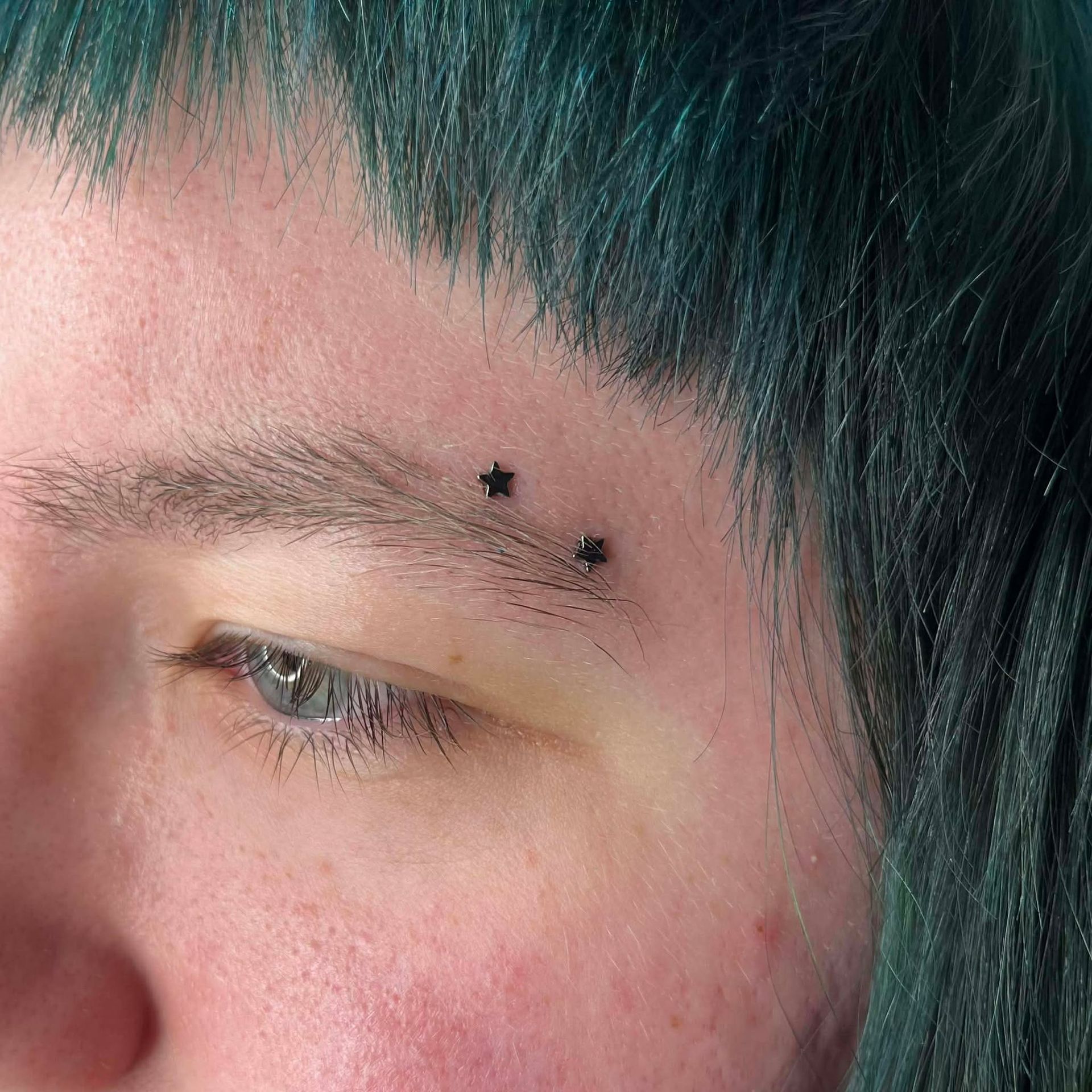 Person's eye and eyebrow with two star-shaped eyebrow piercings and blue hair.